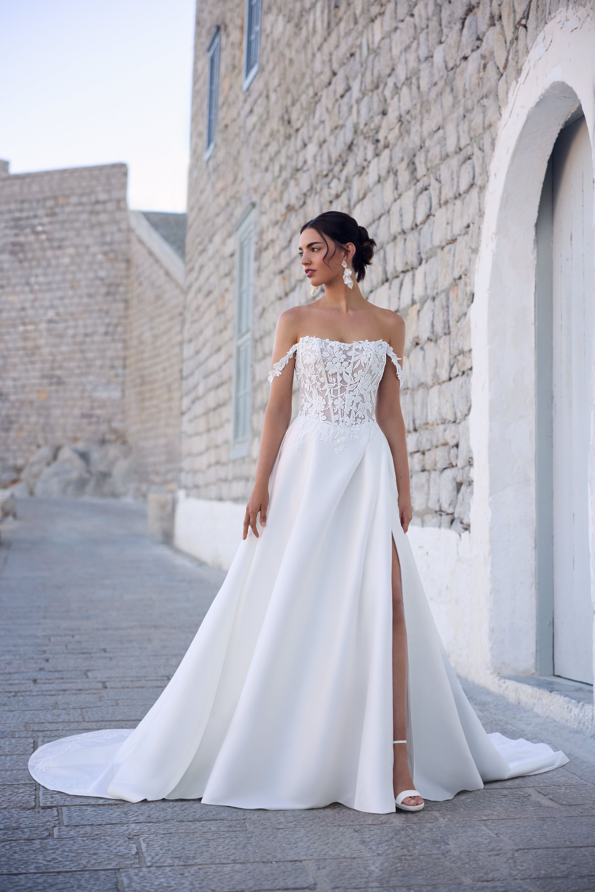 Woman in white strapless wedding dress with slit, standing near stone building.