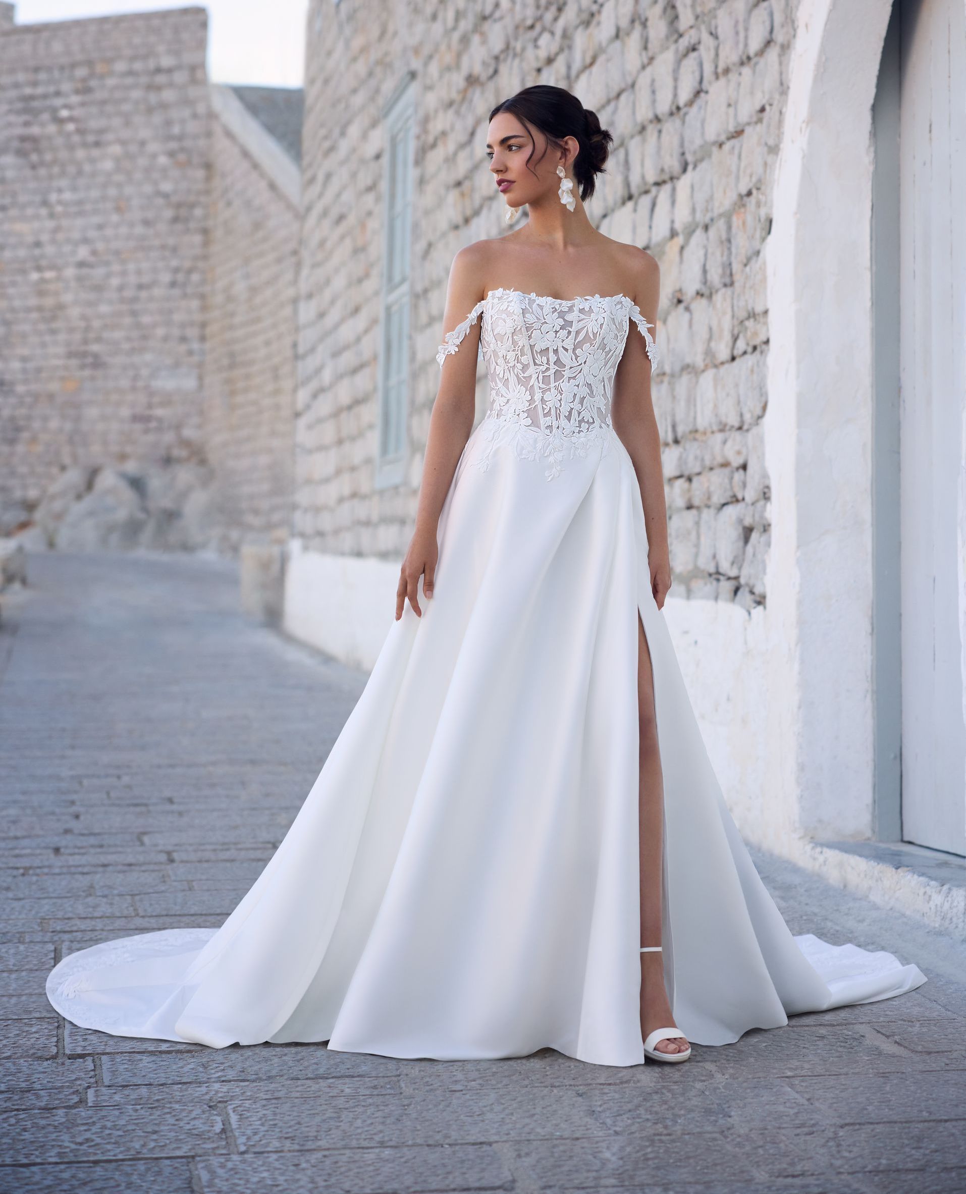 Woman in a white off-the-shoulder wedding dress with a high slit, standing outdoors.