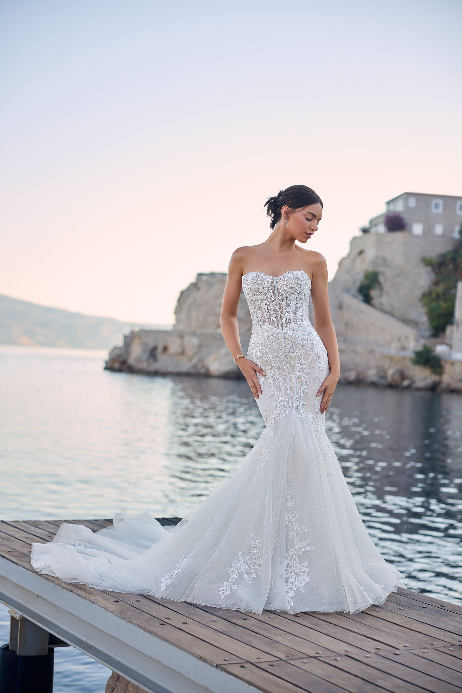 Woman in strapless white wedding dress on wooden dock, ocean and cliffs in the background.