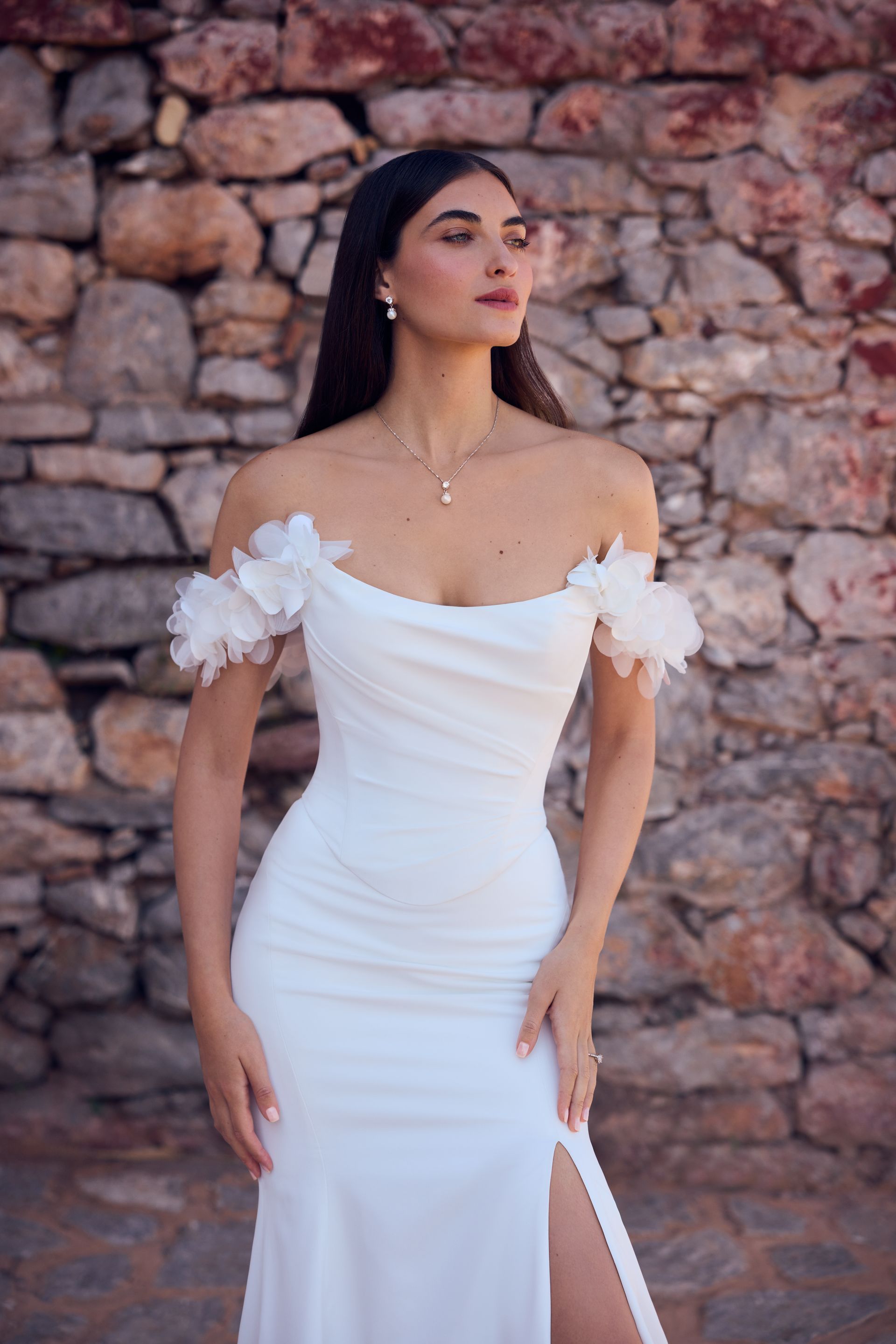 Woman in white off-shoulder gown with floral accents, posing against a stone wall.