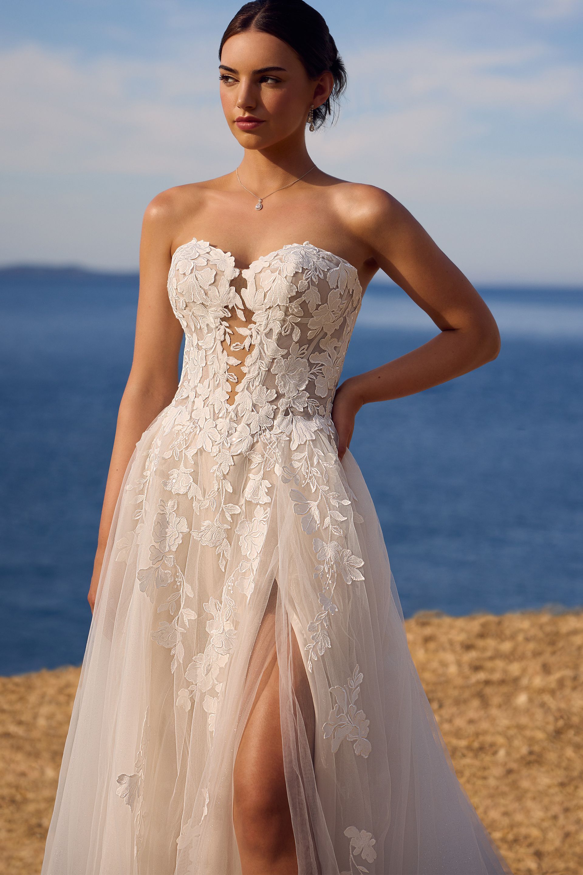 Woman in a strapless white wedding dress with floral embellishments and a high slit, standing by the ocean.