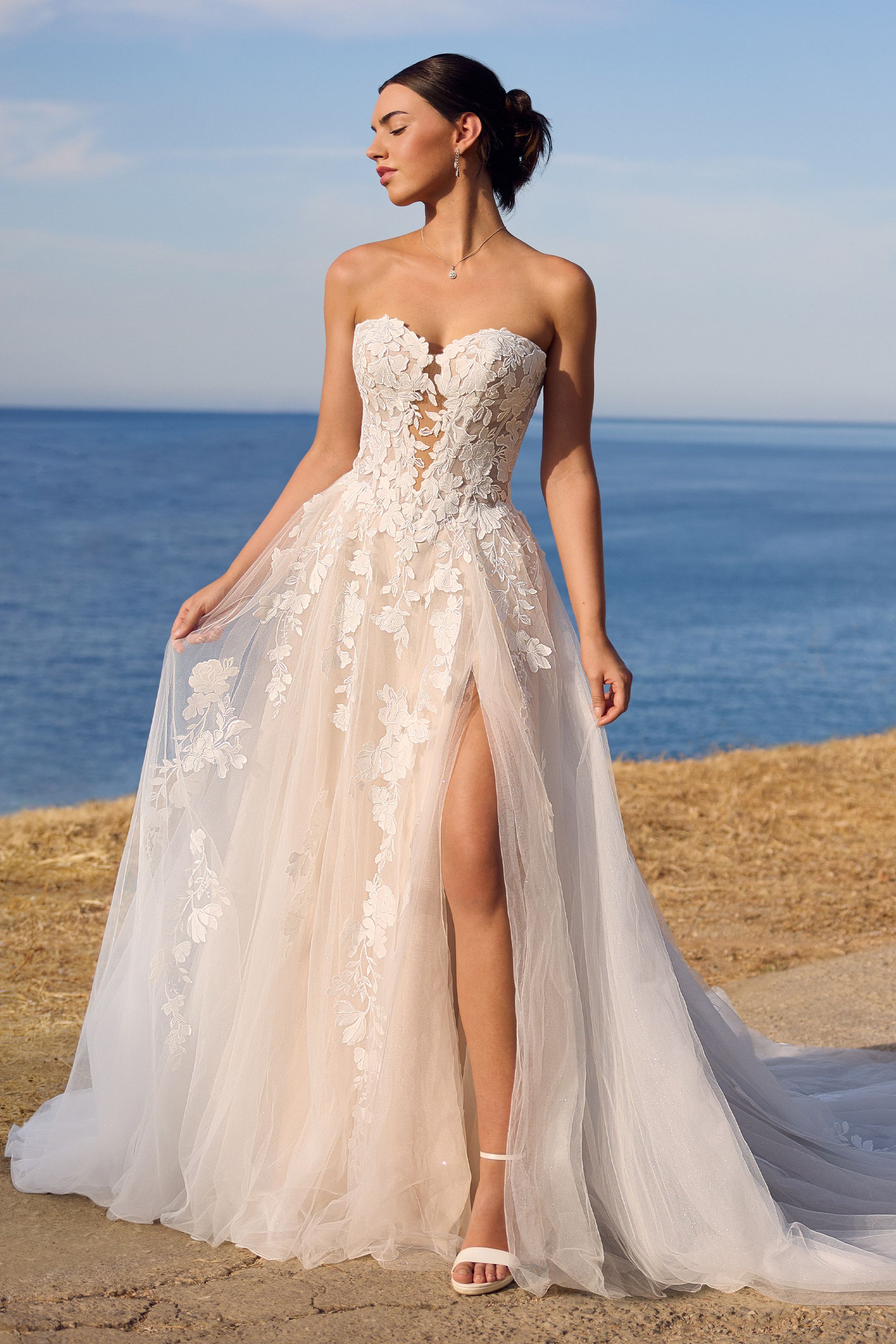 Woman in a strapless white wedding dress with floral embellishments and a high slit, standing by the ocean.