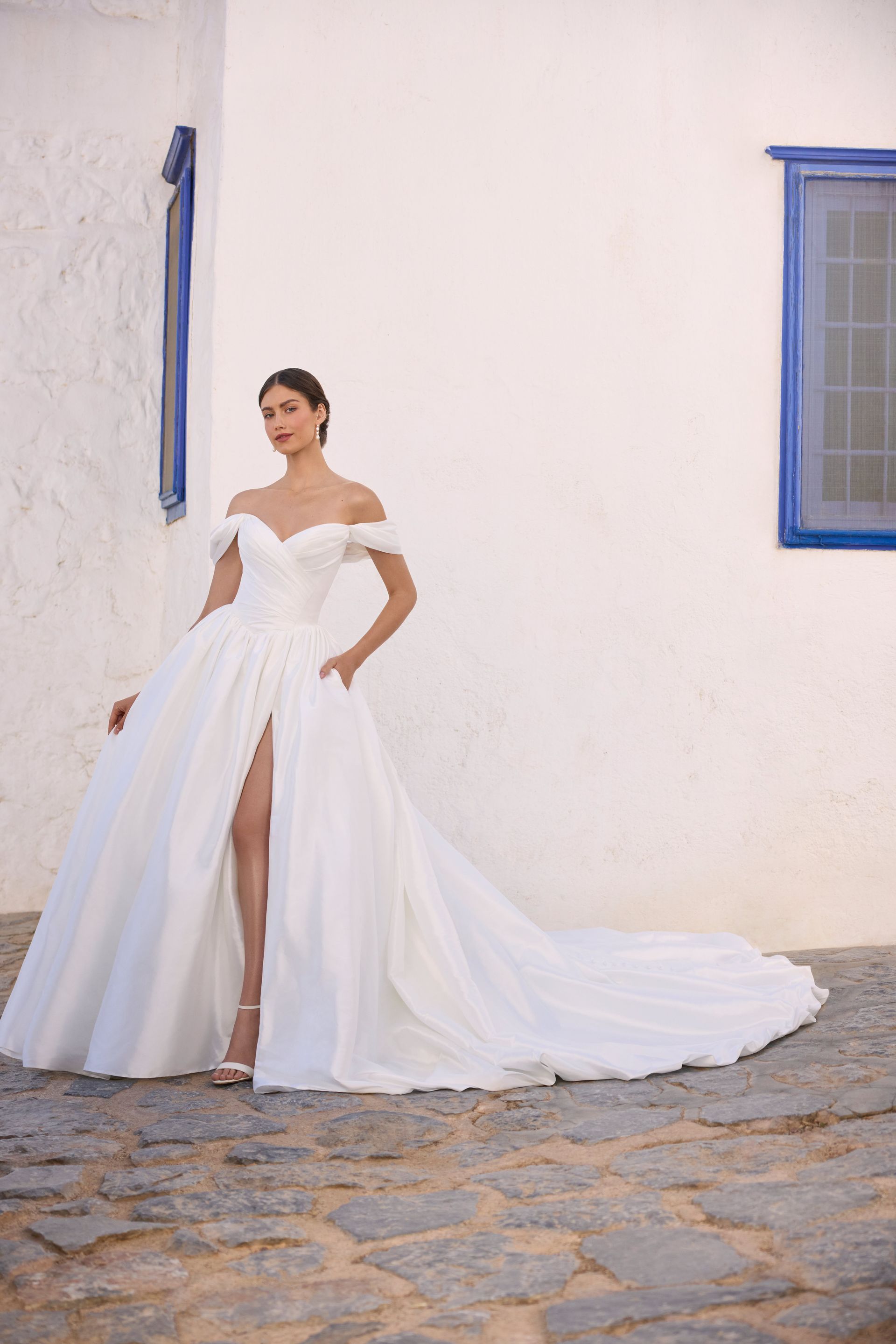 Woman in white off-the-shoulder wedding dress with high slit and long train, standing outside.