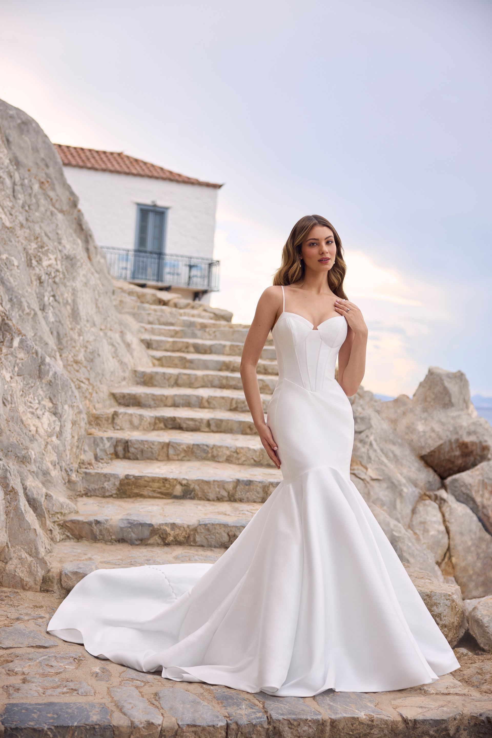 Woman in a white mermaid wedding dress poses on stone steps by a building and the sea.