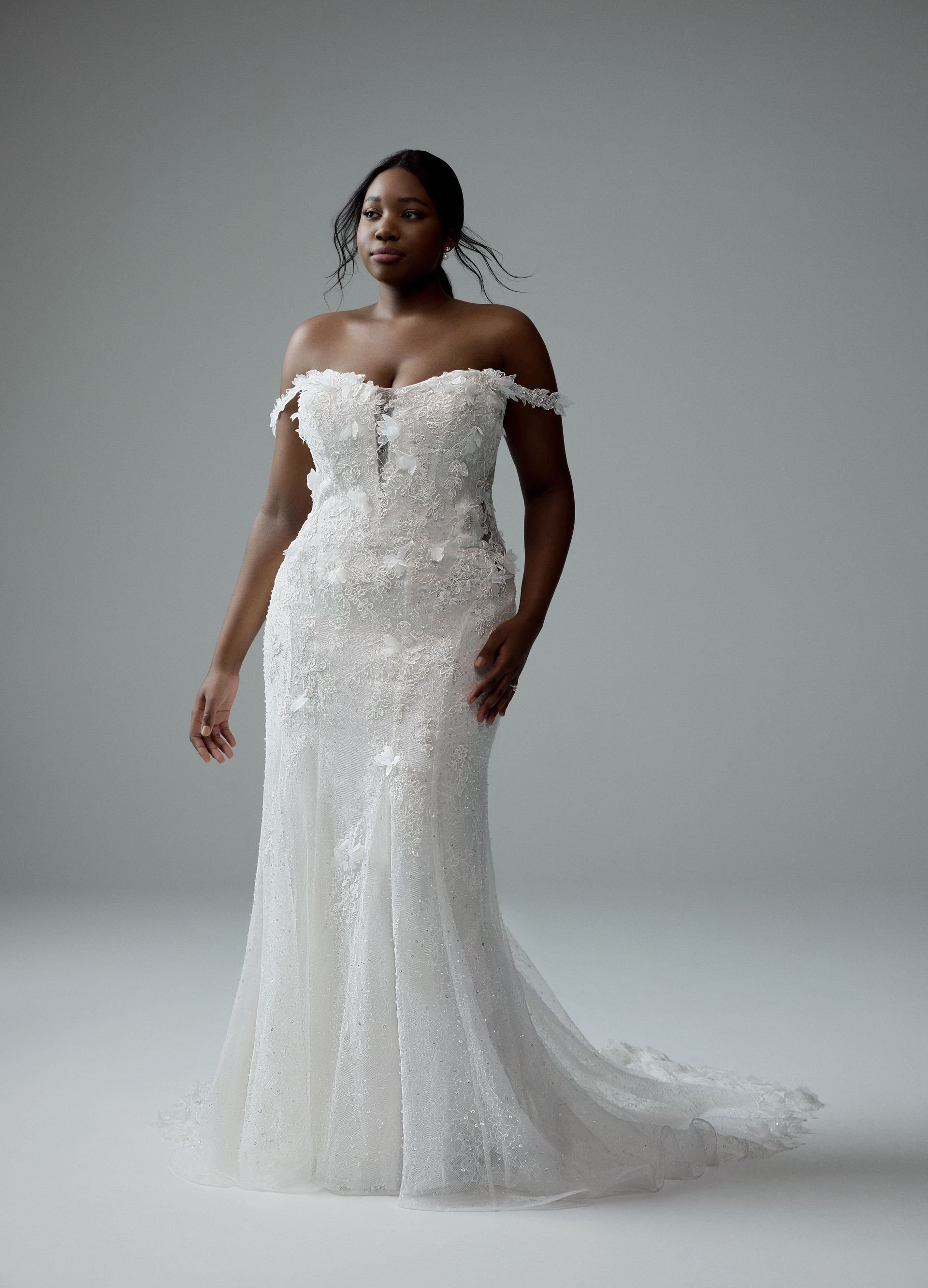 Woman in off-shoulder white wedding dress with floral embellishments; studio shot.