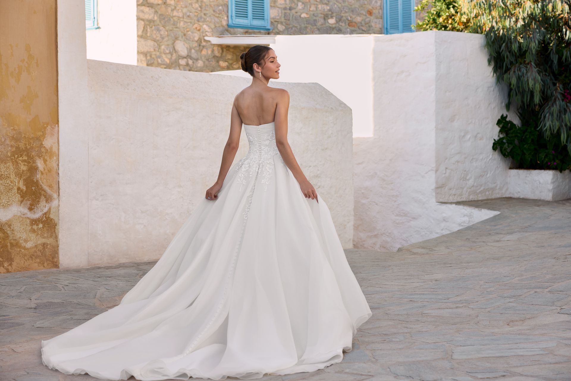 Woman in white strapless wedding gown, turned away, outdoors near white walls.