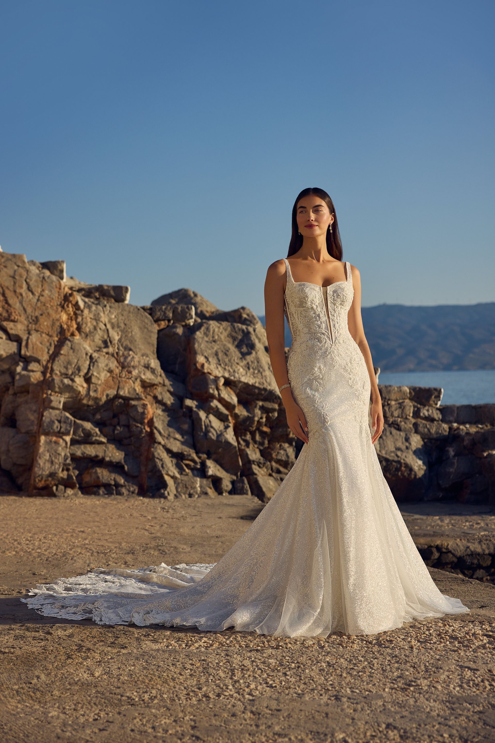 Woman in a beaded mermaid wedding dress stands near rocky coast with a train.