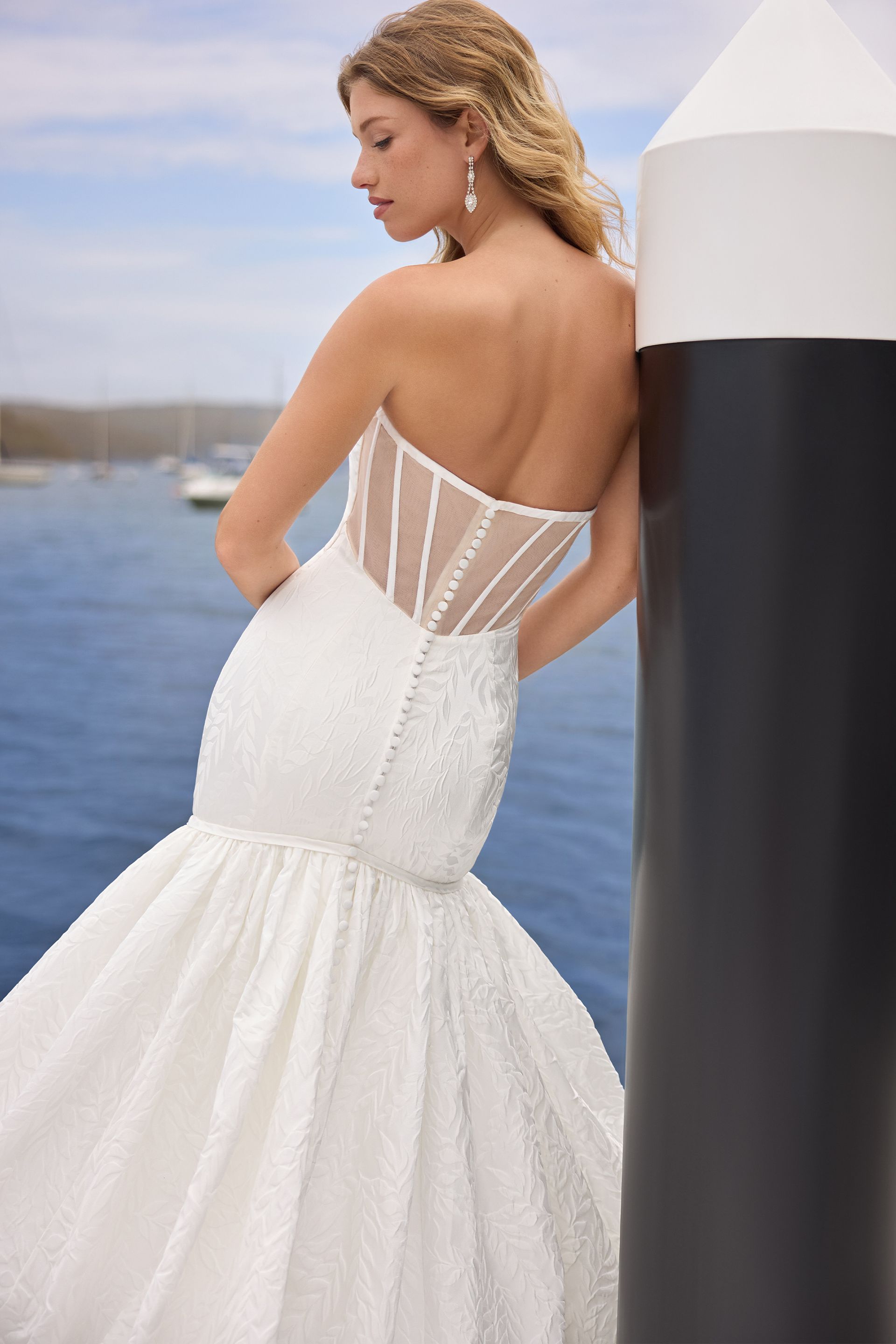 Woman in white strapless wedding gown leans on a black and white post near water.