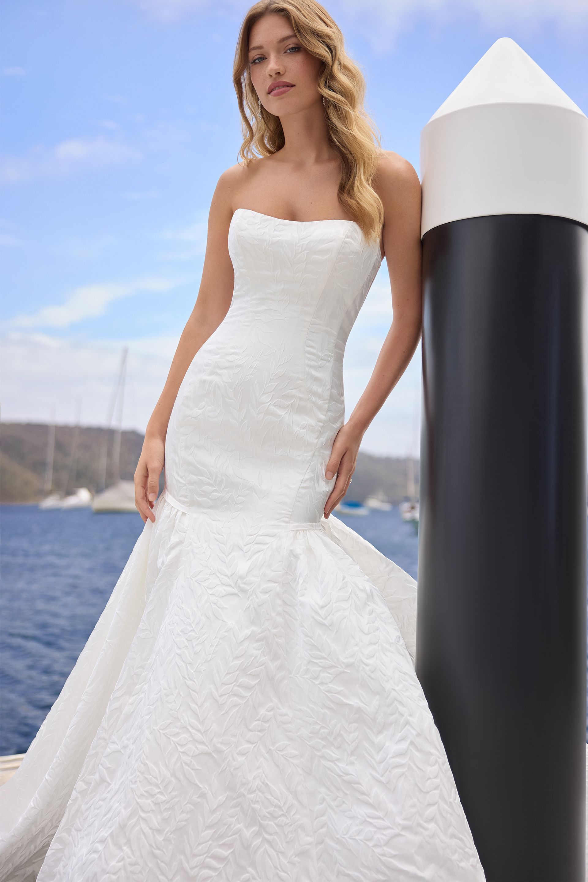 Woman in white strapless wedding gown leans on a black and white post near water.
