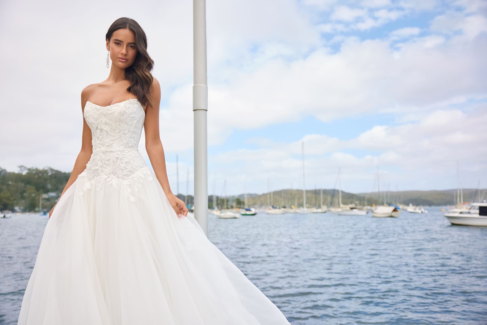 Woman in wedding dress stands on pier, water and boats in background.