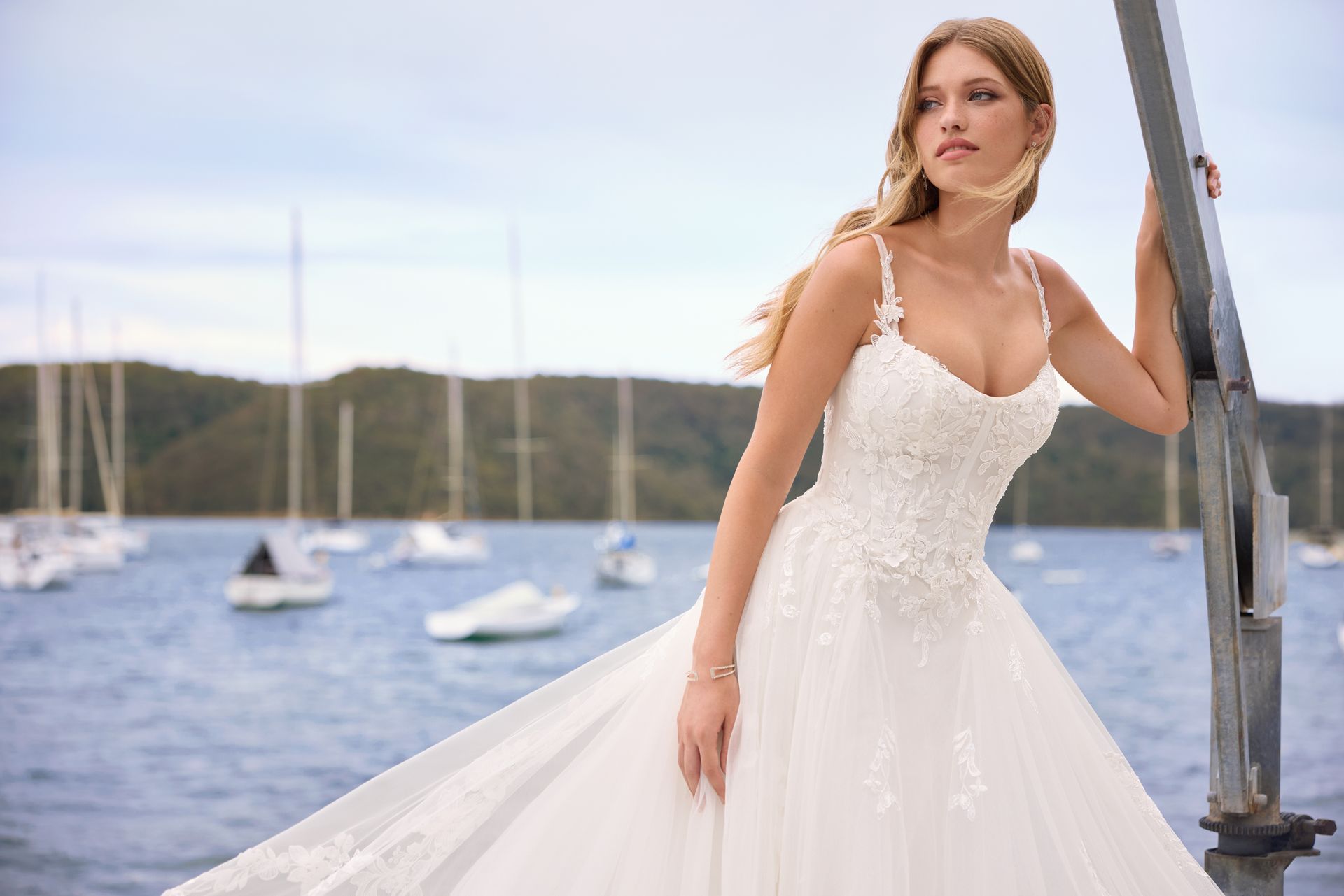 Woman in a white wedding gown, leaning on a metal pole near a body of water with sailboats.