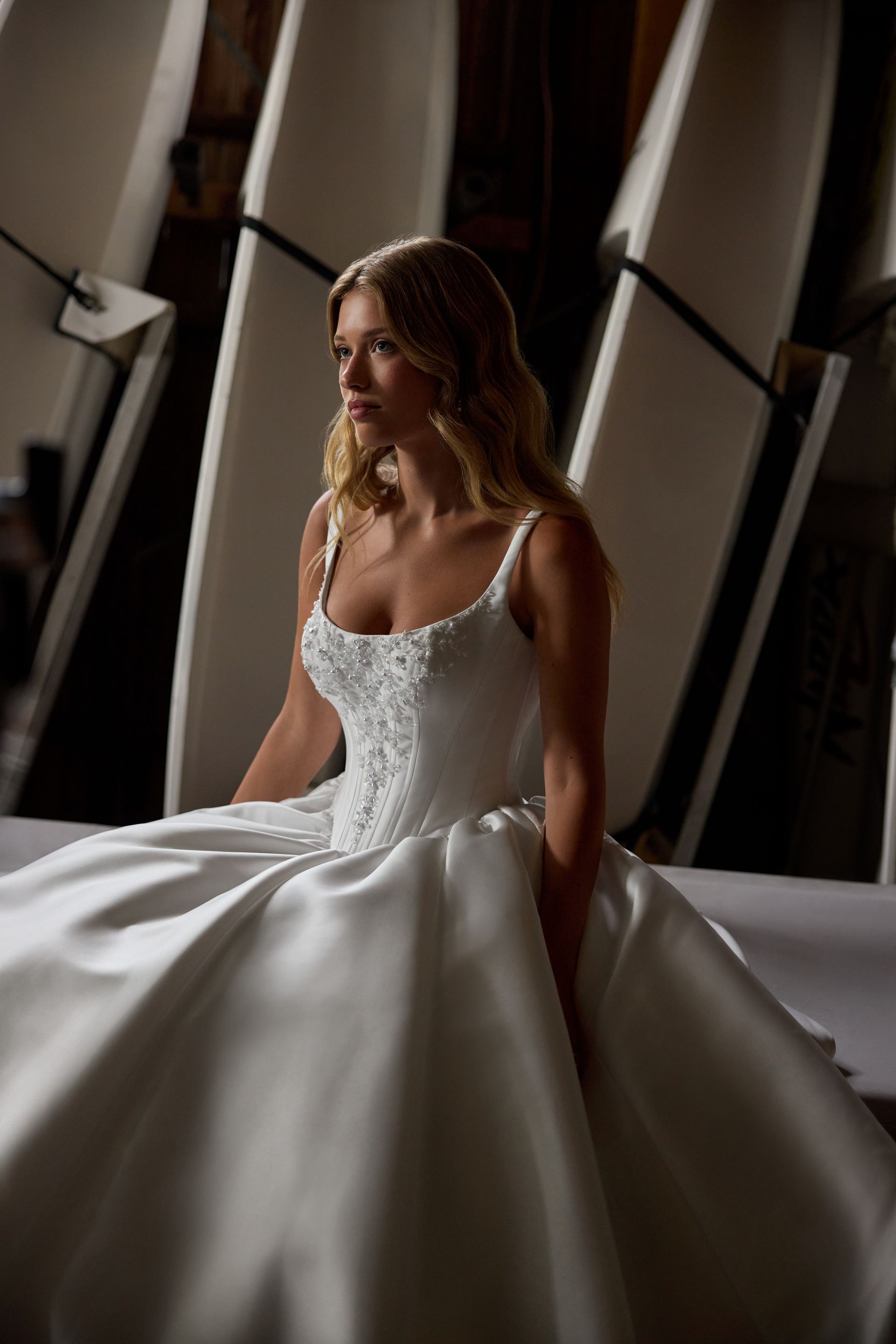 Woman in a white wedding dress, looking away, seated in front of white structures.