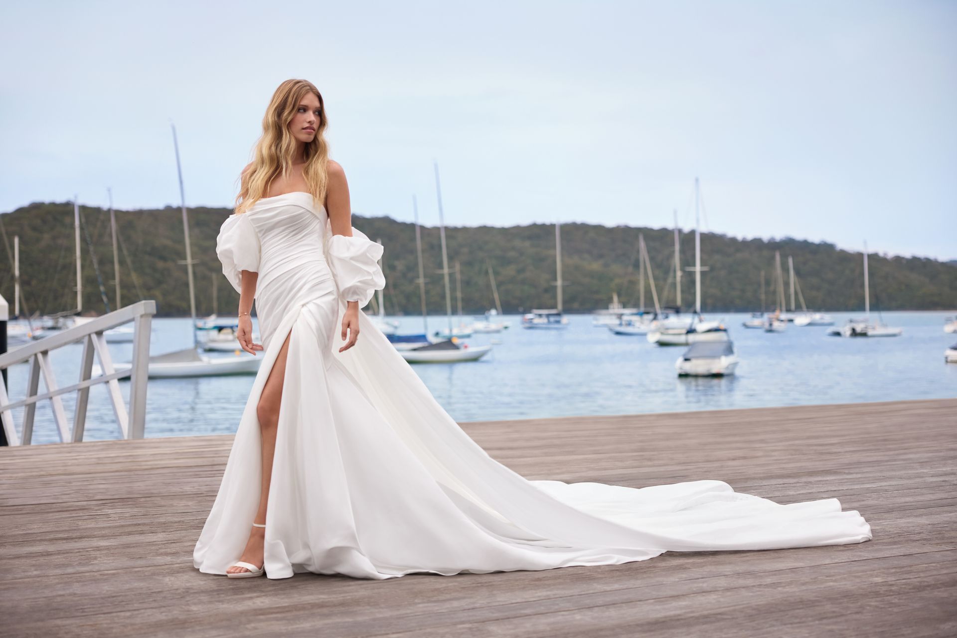 Woman in white wedding dress with train, standing on a pier overlooking boats and water.