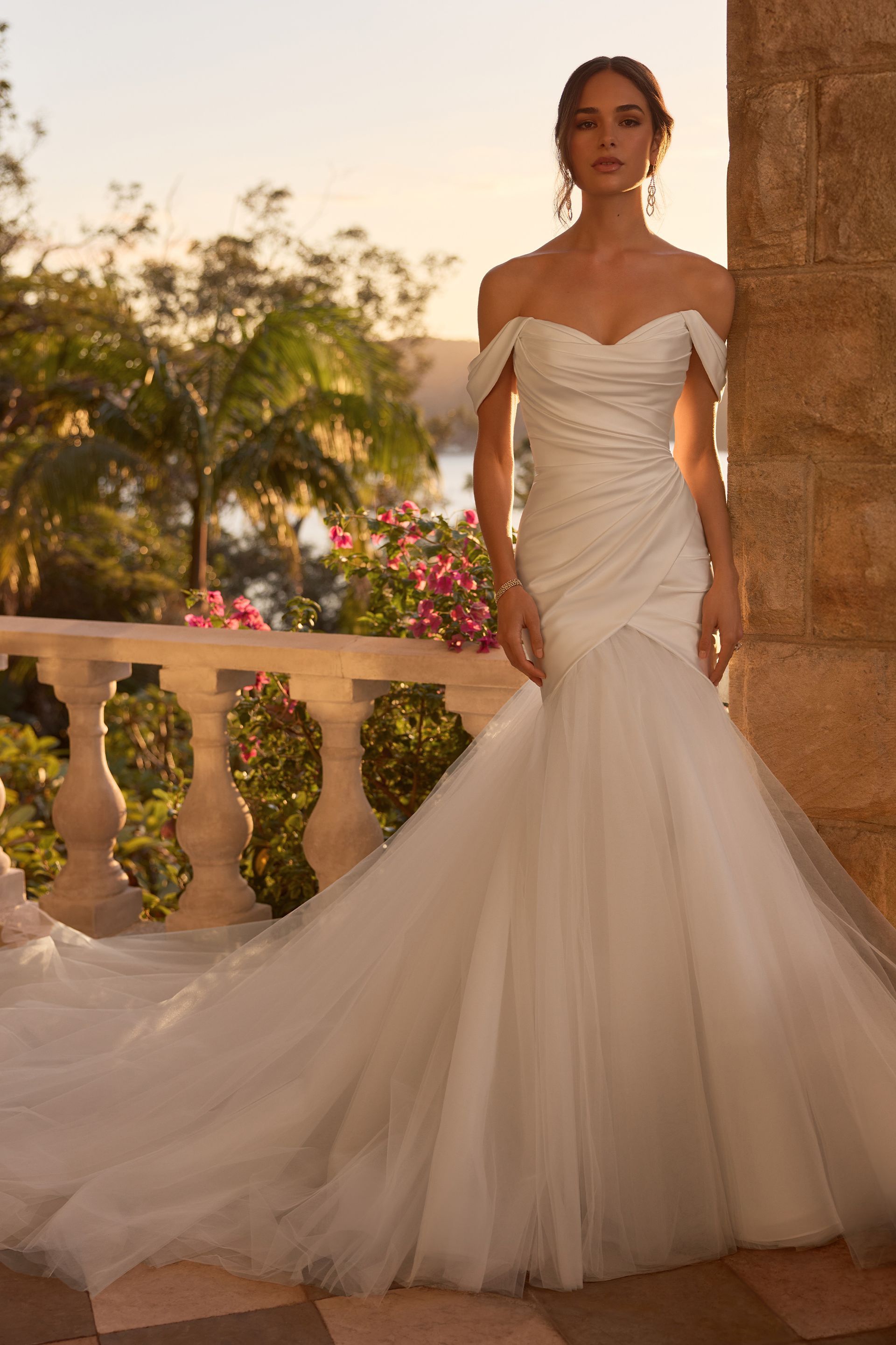 Woman in a white off-the-shoulder wedding gown with a long train, standing on a balcony overlooking water at sunset.