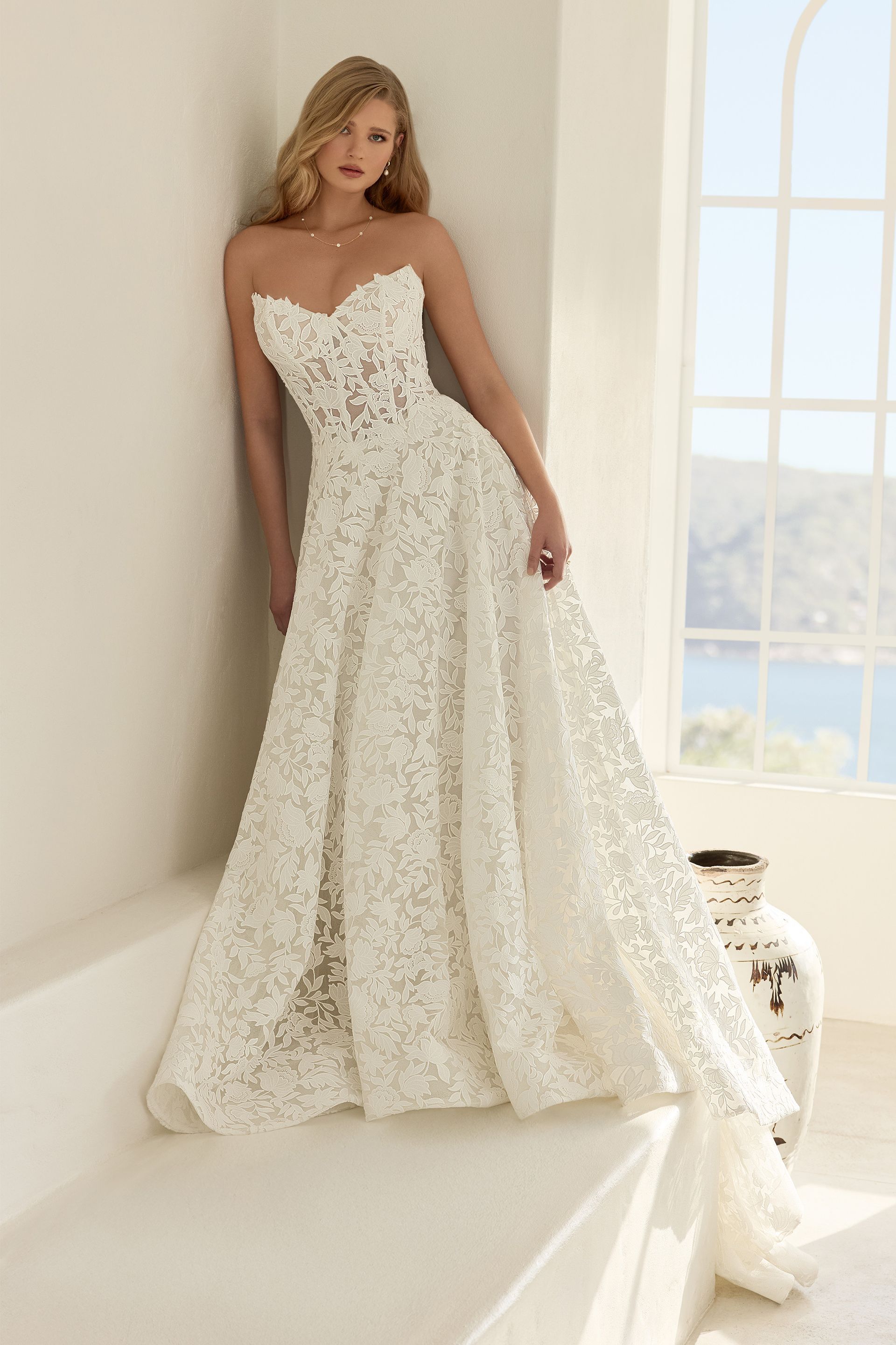 Woman in white strapless wedding gown with floral embellishments, leaning against white wall near window, overlooking ocean.