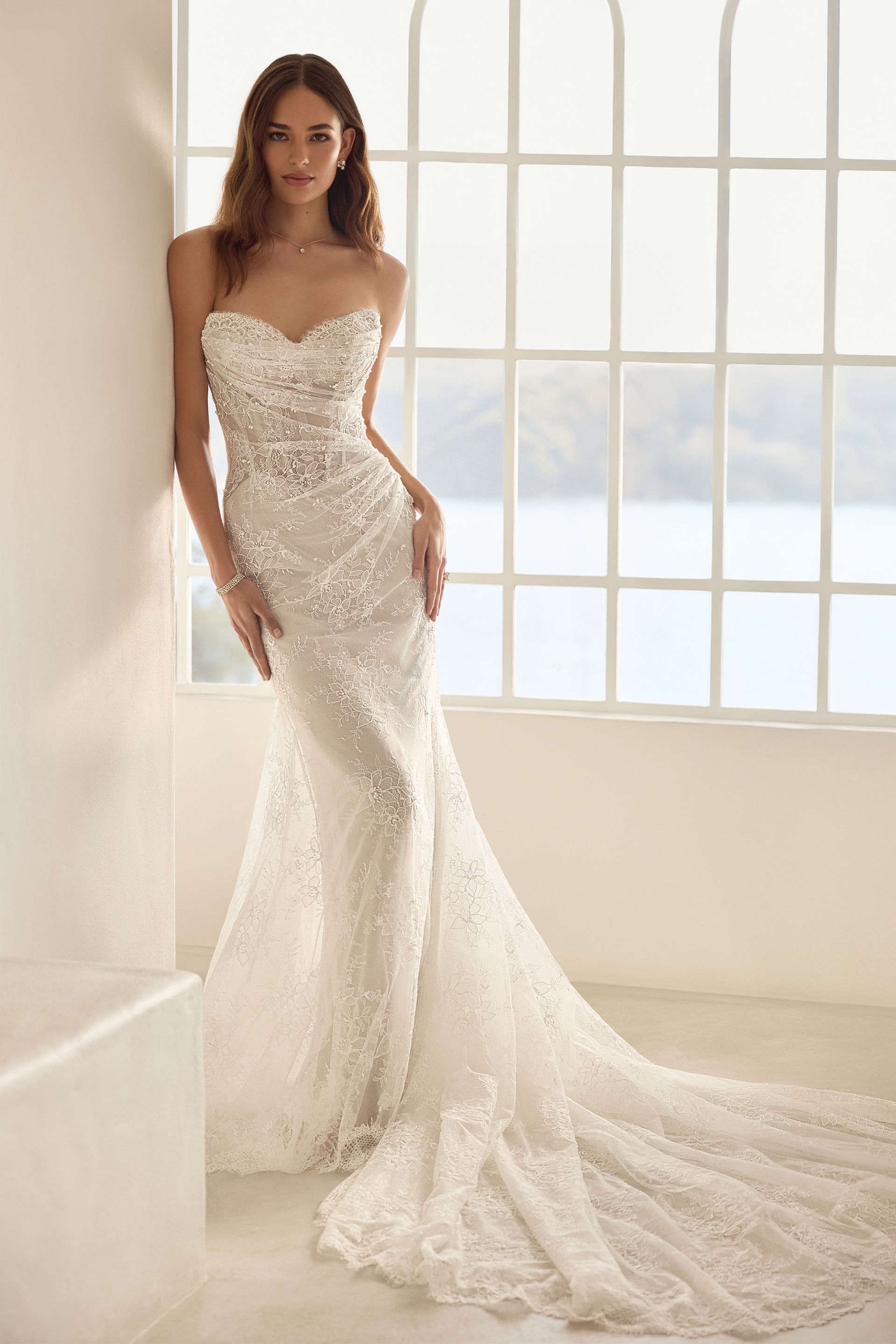 Woman in a strapless lace wedding gown standing by a large window in a bright room