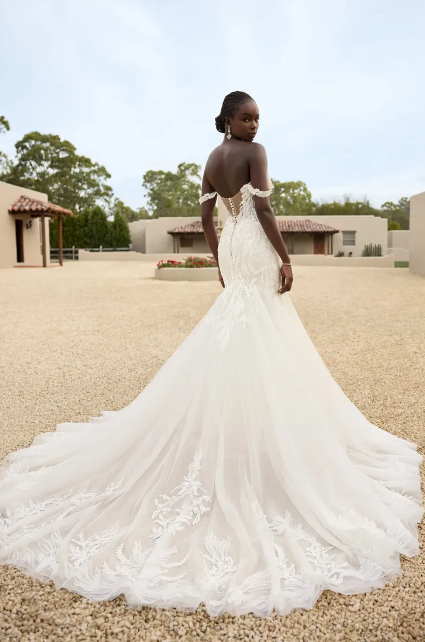 Woman in an off-the-shoulder wedding dress with a long train, posing outside.