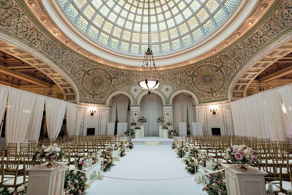 Elegant wedding ceremony setting. White aisle, floral arrangements, gold chairs, under a domed ceiling.