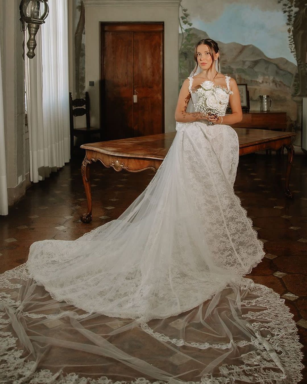 Woman in white lace wedding dress with long train, holding bouquet indoors.