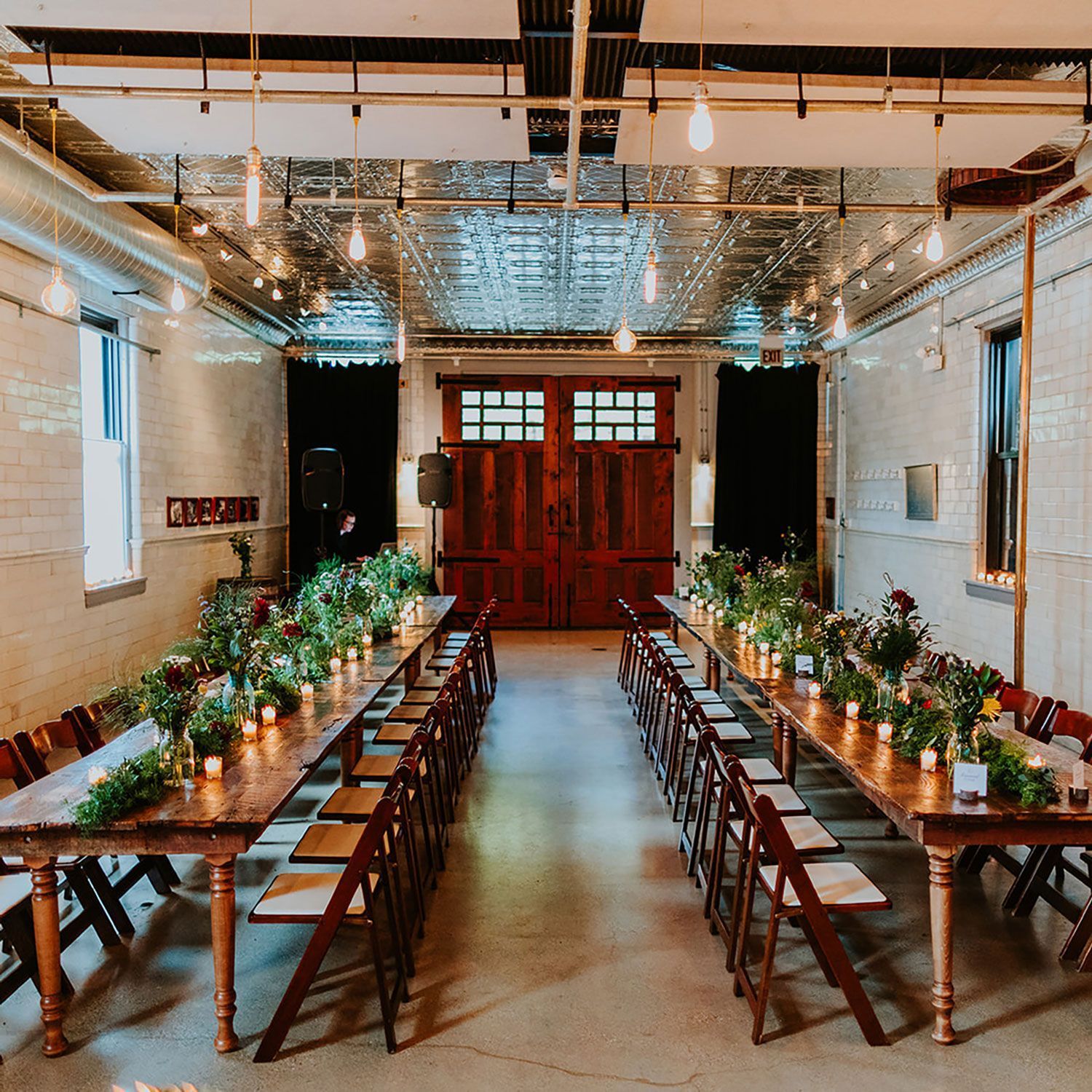 Long wooden tables set for a banquet in a rustic room with exposed brick, strung lights, and floral centerpieces.