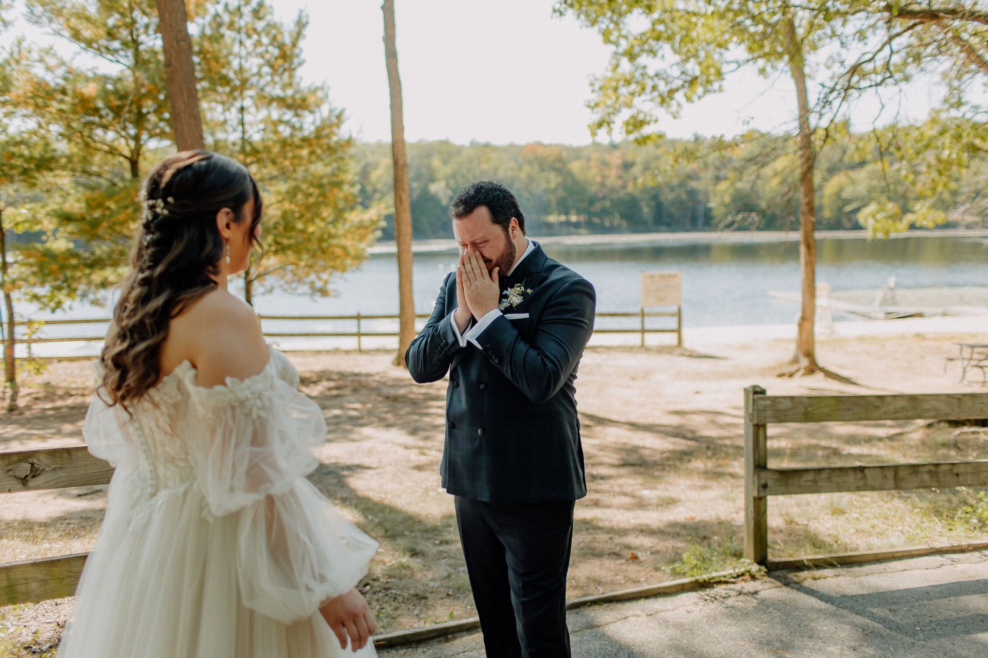 Groom wiping tears during a first look with bride in a forest setting by a lake.
