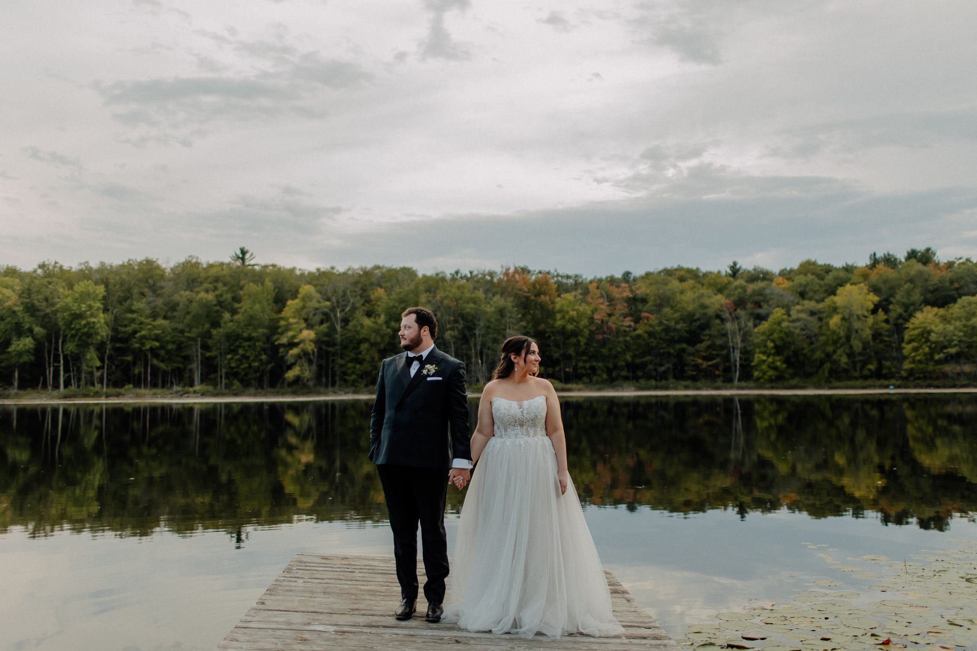 Couple holding hands, standing on a dock, lake and trees in the background, overcast sky.