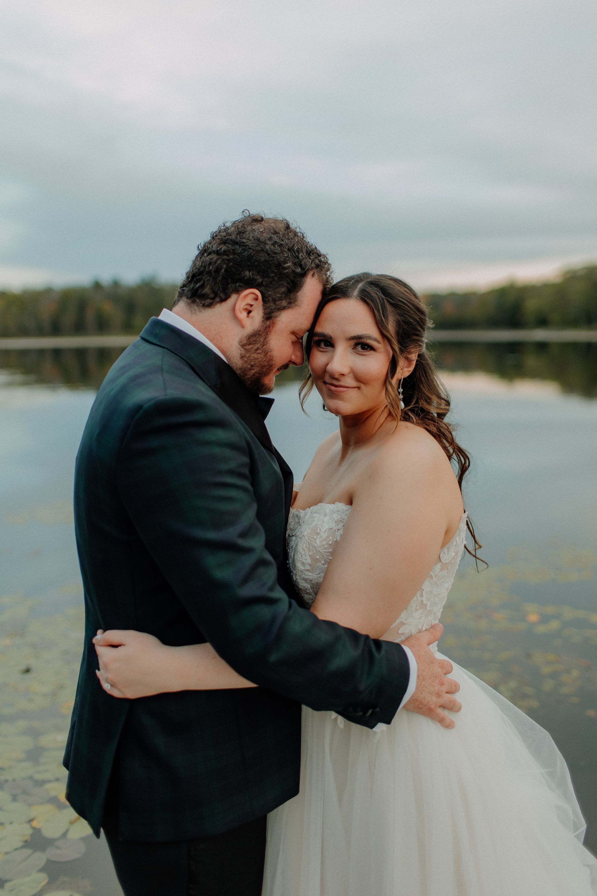 Couple embraces near lake; bride wears strapless white gown, groom in navy suit; cloudy sky.