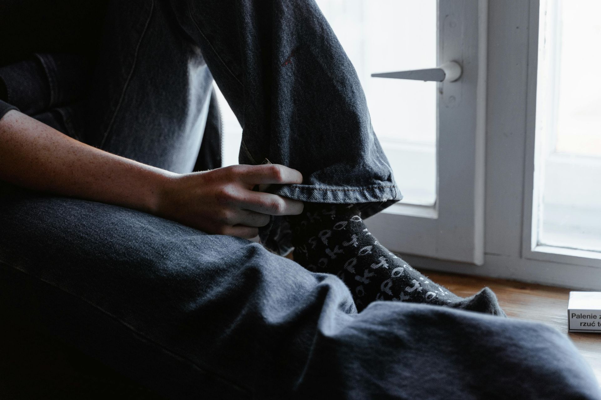 Person sits on windowsill, holding edge of pants. Gray pants, patterned socks, window. Person sits on windowsill, holding edge of pants. Gray pants, patterned socks, window.