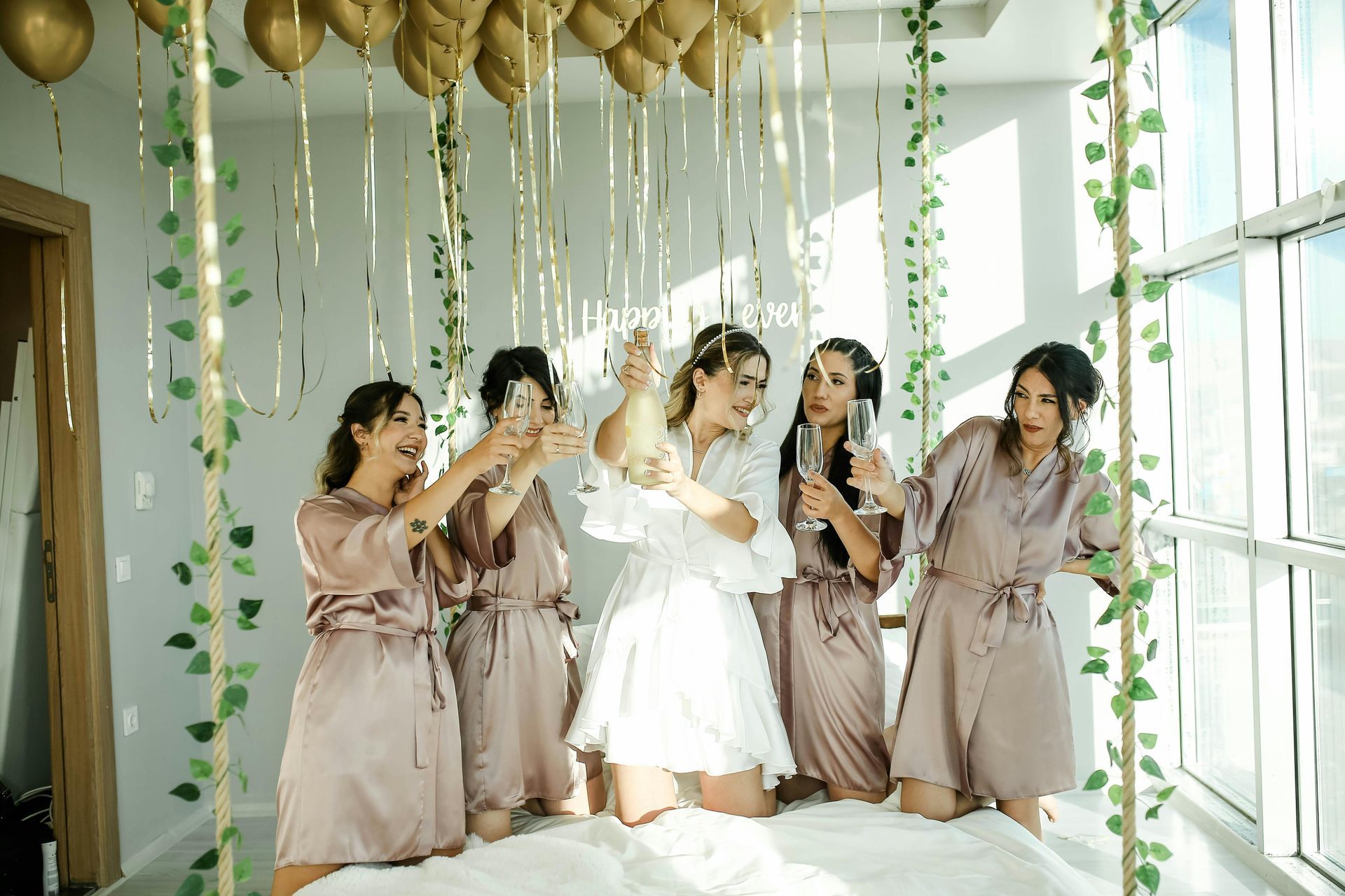 Women celebrating with champagne, wearing robes, inside a room decorated with balloons and vines. Women celebrating with champagne, wearing robes, inside a room decorated with balloons and vines.