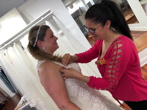 Woman in white dress being fitted by another woman in a red shirt at a bridal shop.