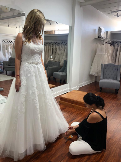 Bride in a white wedding dress being fitted by a person kneeling on the floor in a bridal shop.