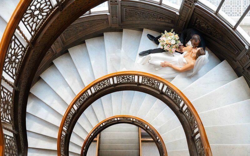 Bride and groom on a spiral staircase, looking at each other. White dress, holding flowers, ornate wood railing.