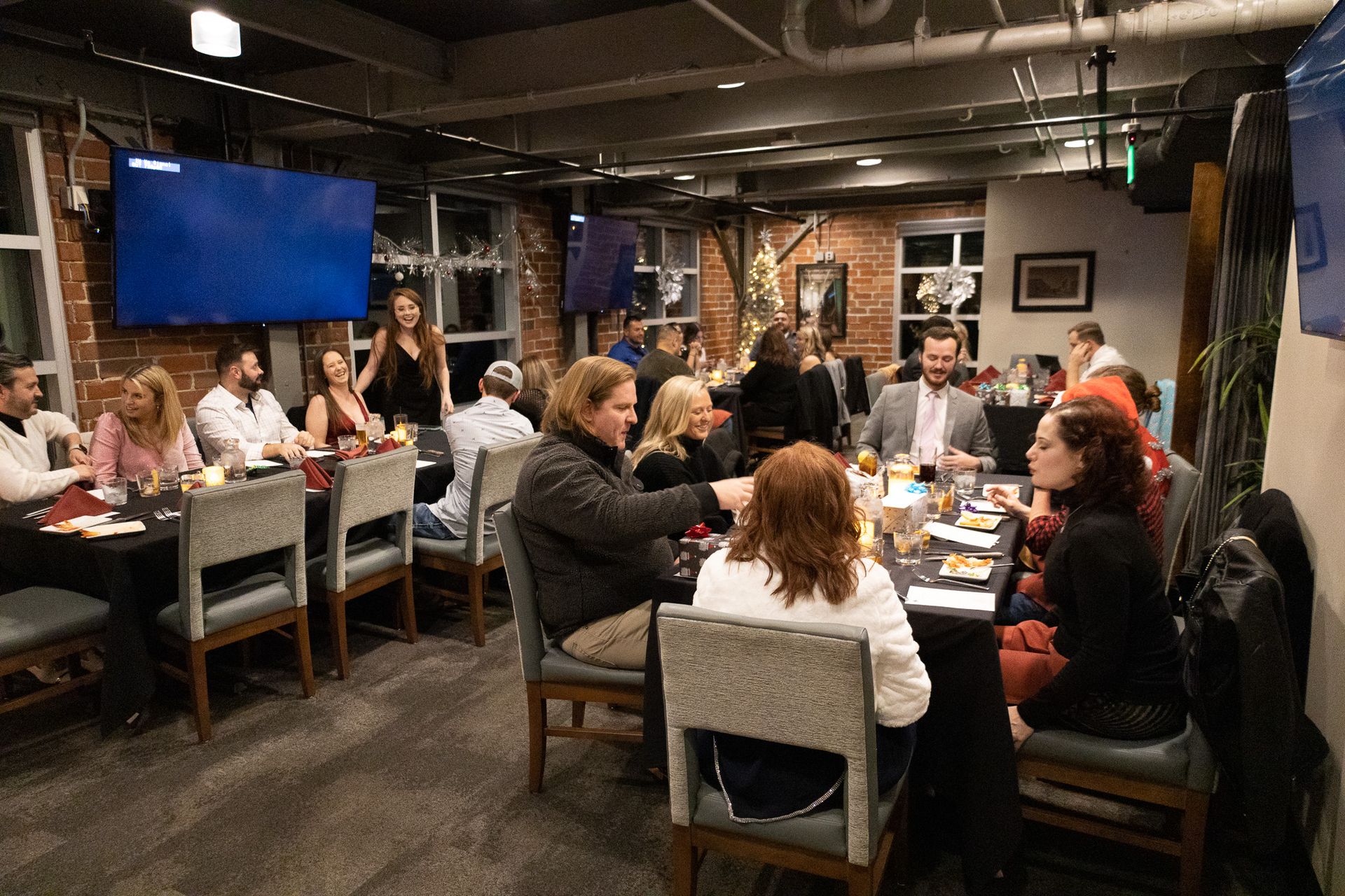 People seated at tables in a dimly lit room, possibly a restaurant. Television screens and brick walls are visible.
