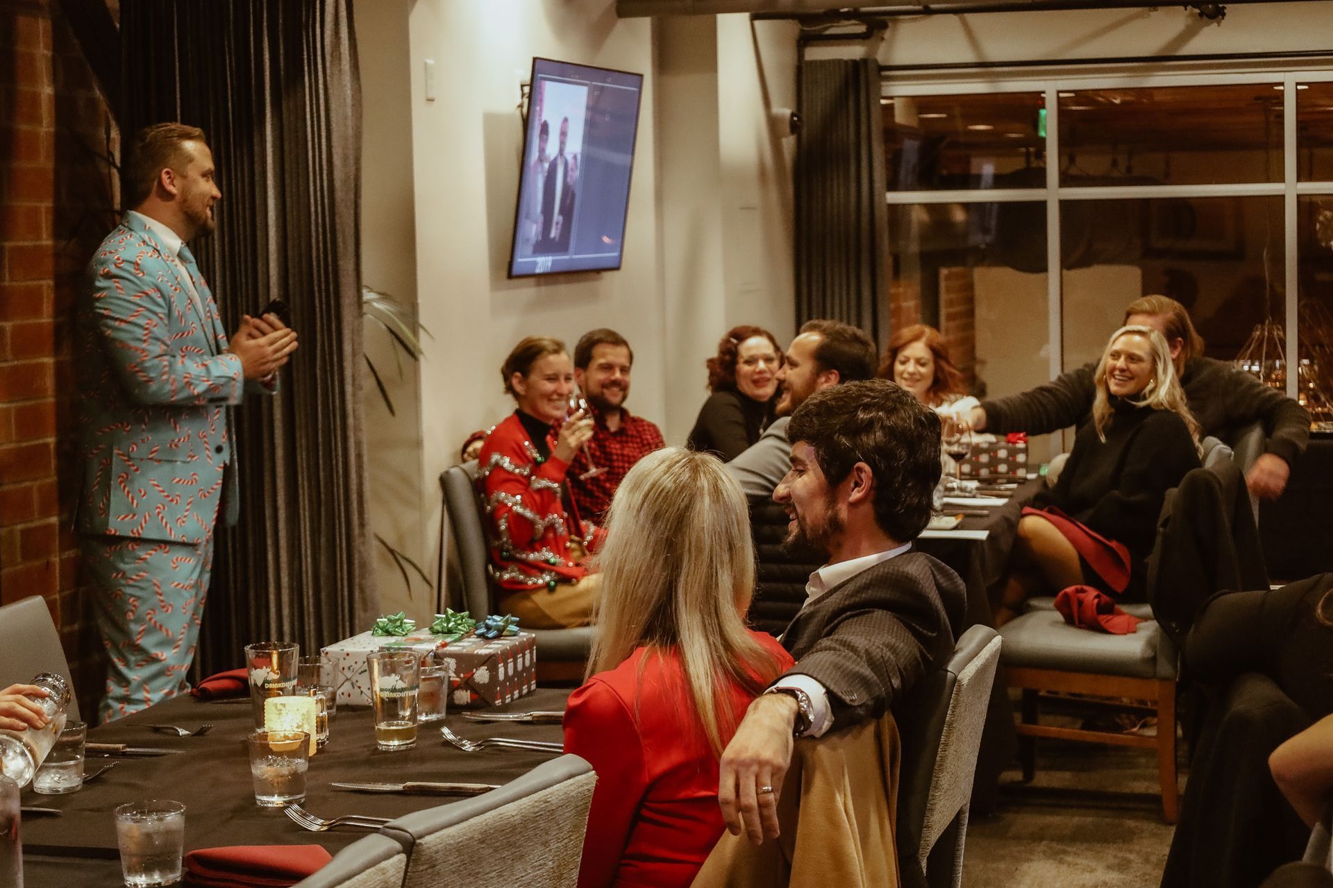 Man in suit speaks to group at table, some laughing. Formal setting, interior.
