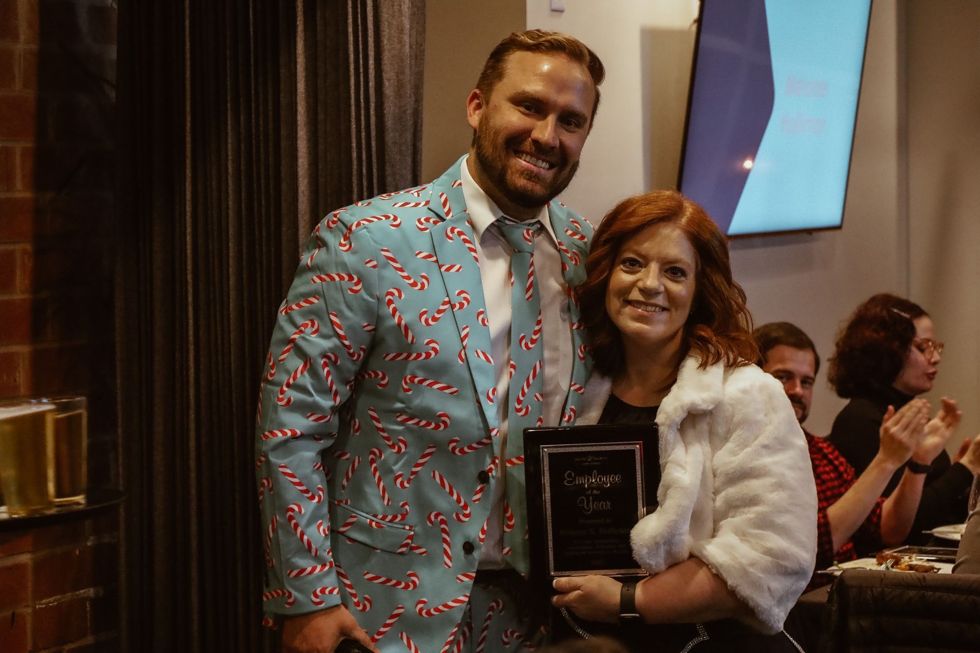 Man in candy cane suit and woman in fur coat pose, holding an award at an event.