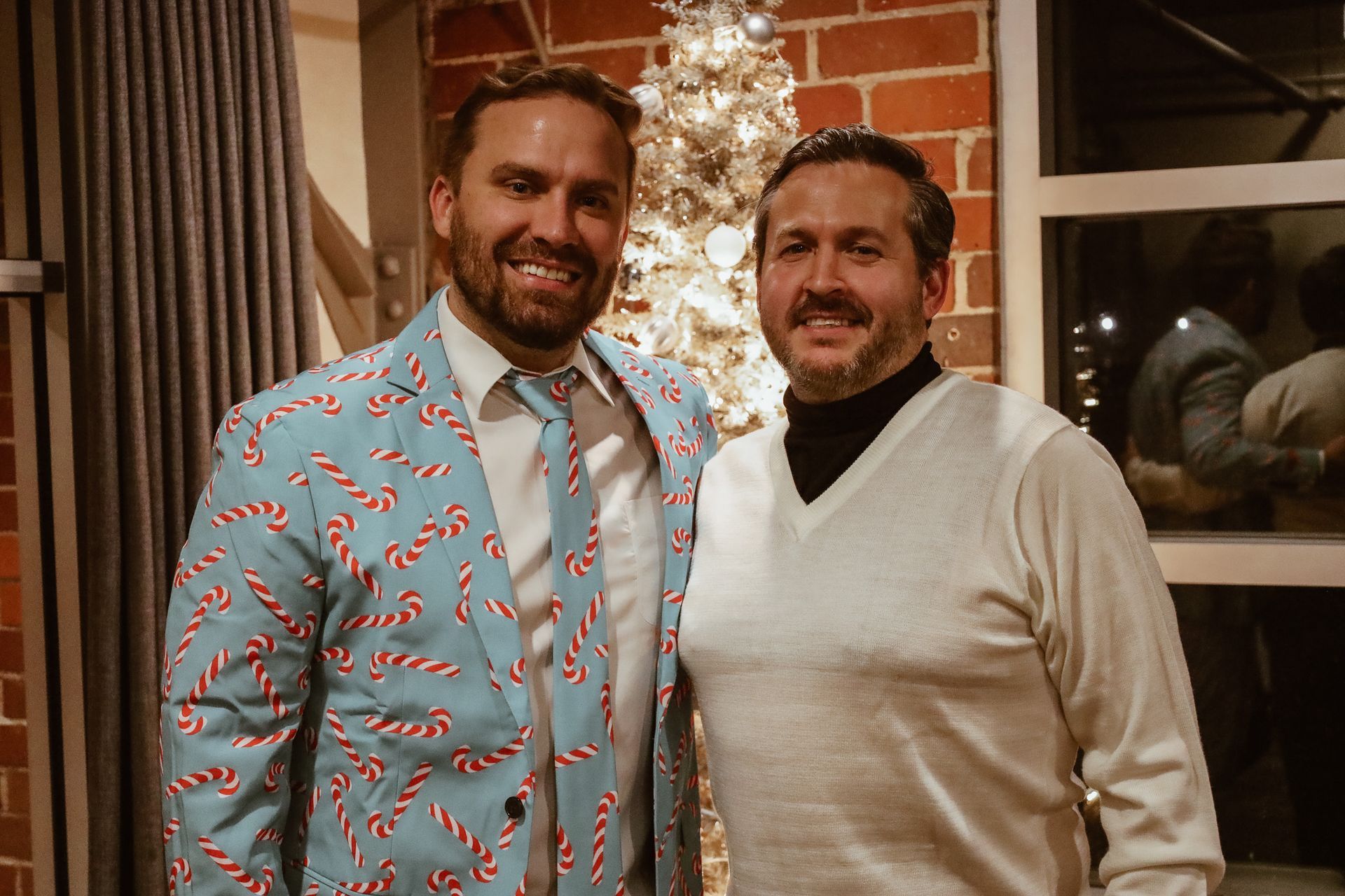 Two men smiling. One in a candy cane suit, other in a white sweater, near a Christmas tree.