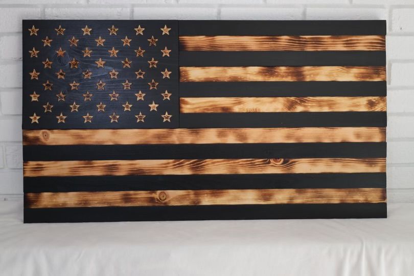Wooden American flag art with charred stripes and star field, on a white brick wall.