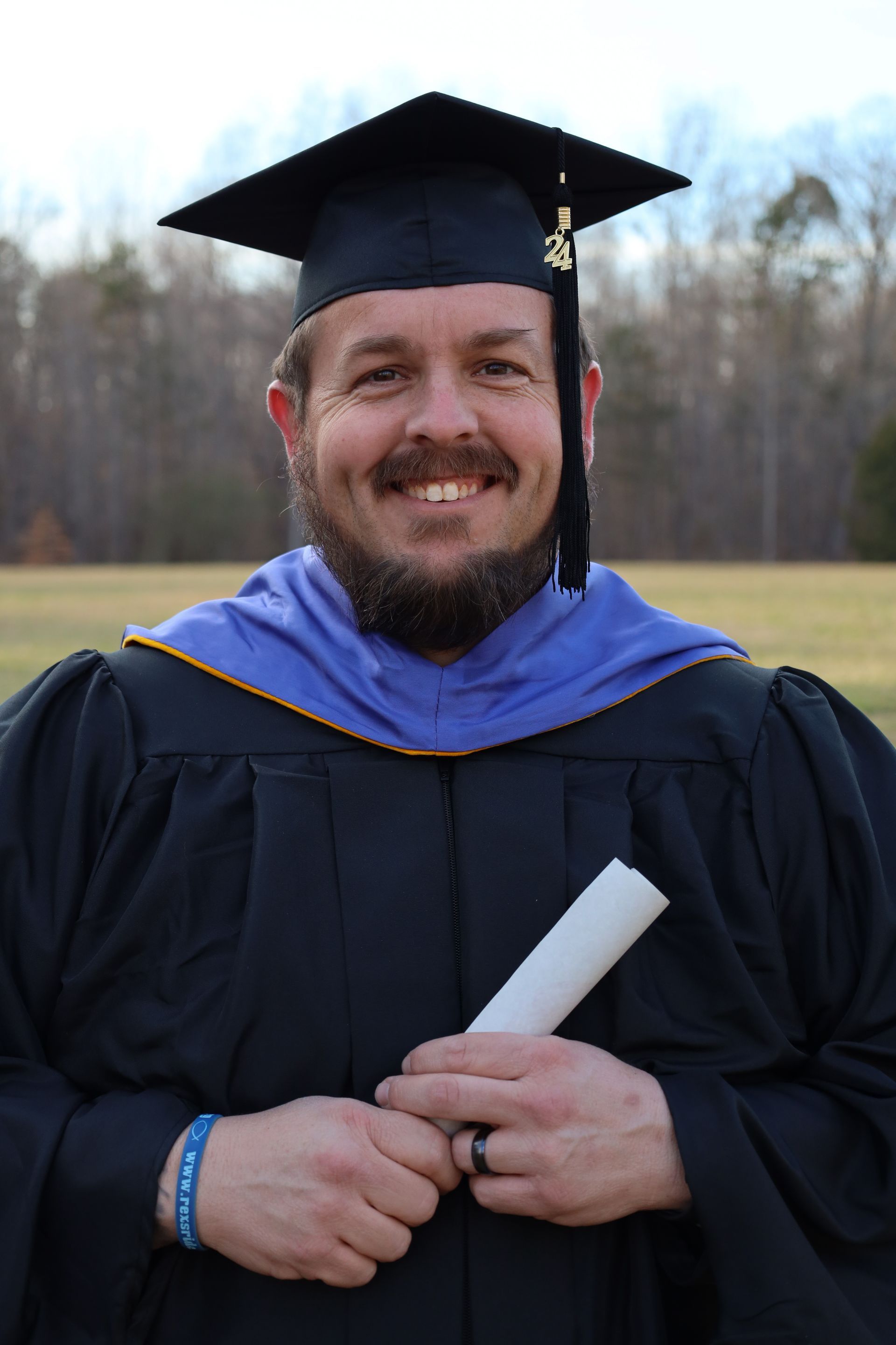 Man in graduation cap and gown smiles, holding diploma, outdoor setting.