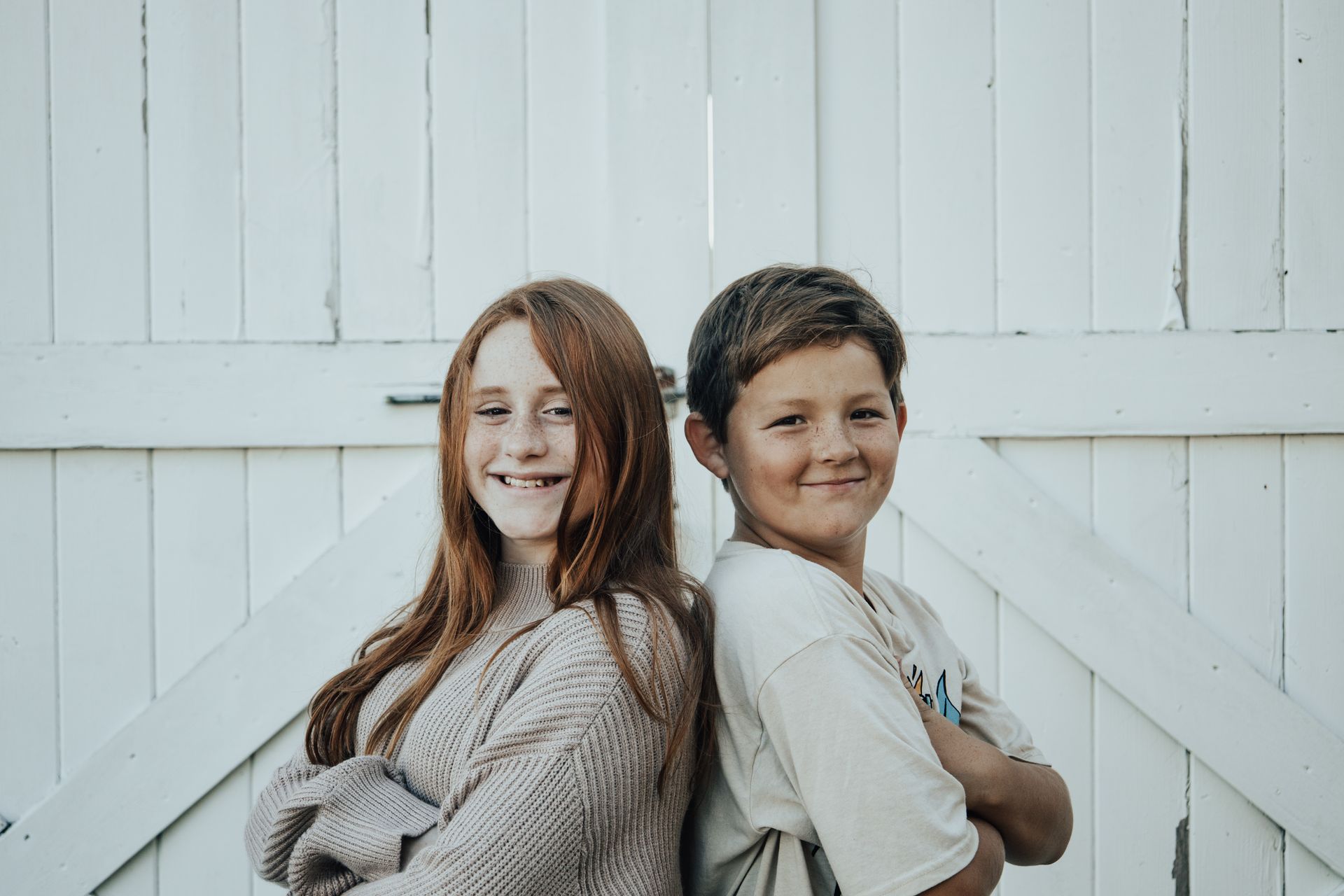 Two children smiling, standing back to back against a white wooden door, arms crossed.