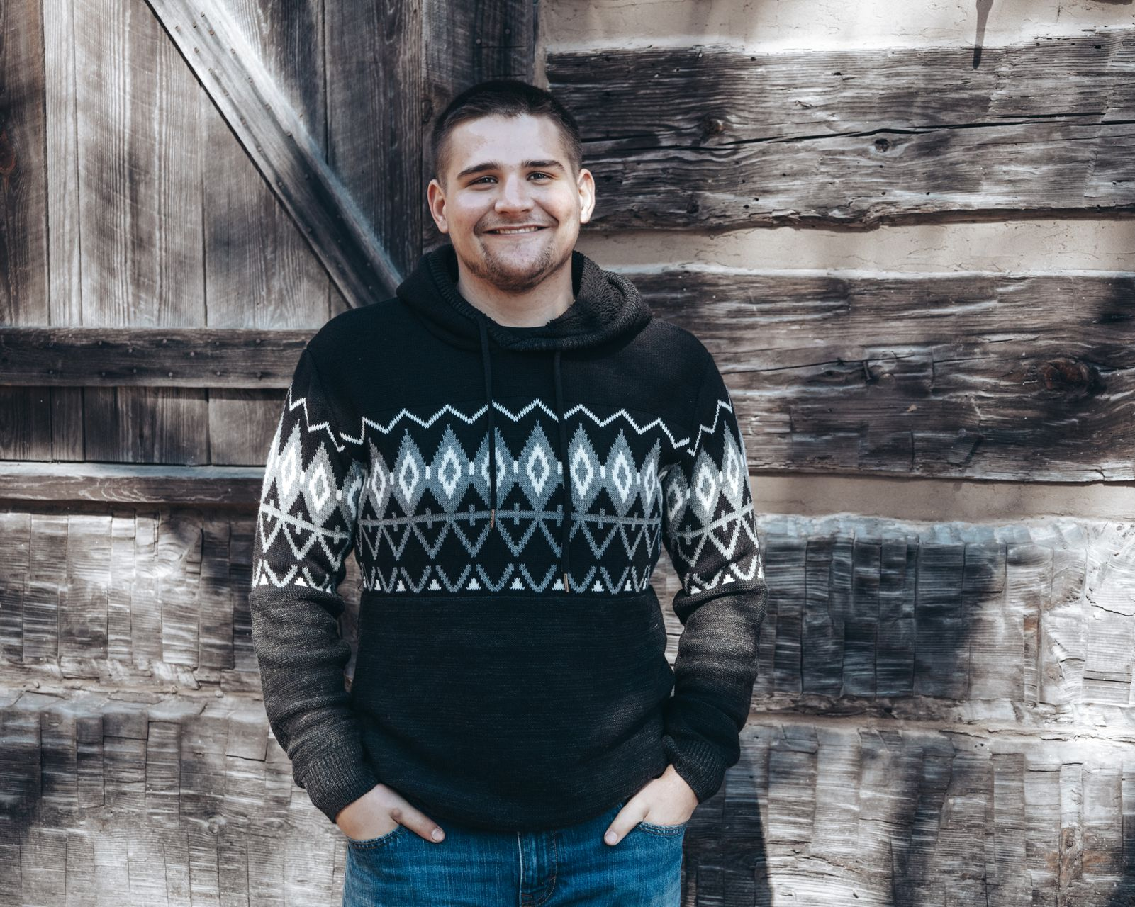 Man in patterned sweater, hands in pockets, smiling in front of weathered wooden wall.