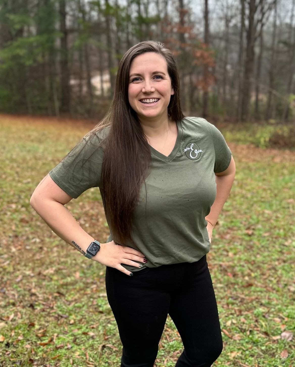 Woman in olive green shirt and black pants, smiling with hands on hips in a grassy outdoor setting.