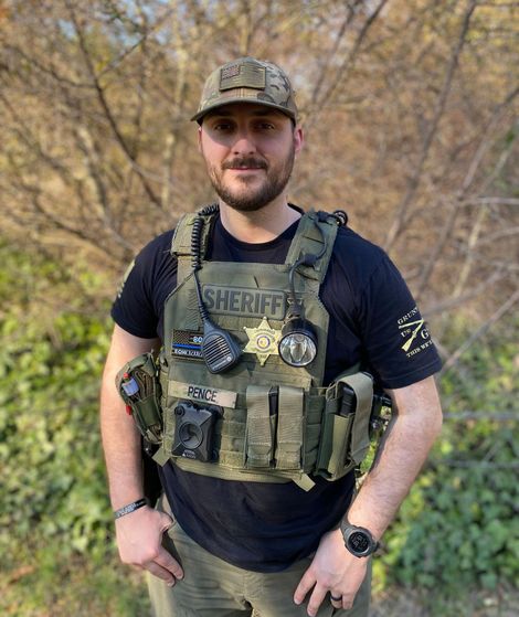 Sheriff in tactical vest and camouflage hat, standing outside.