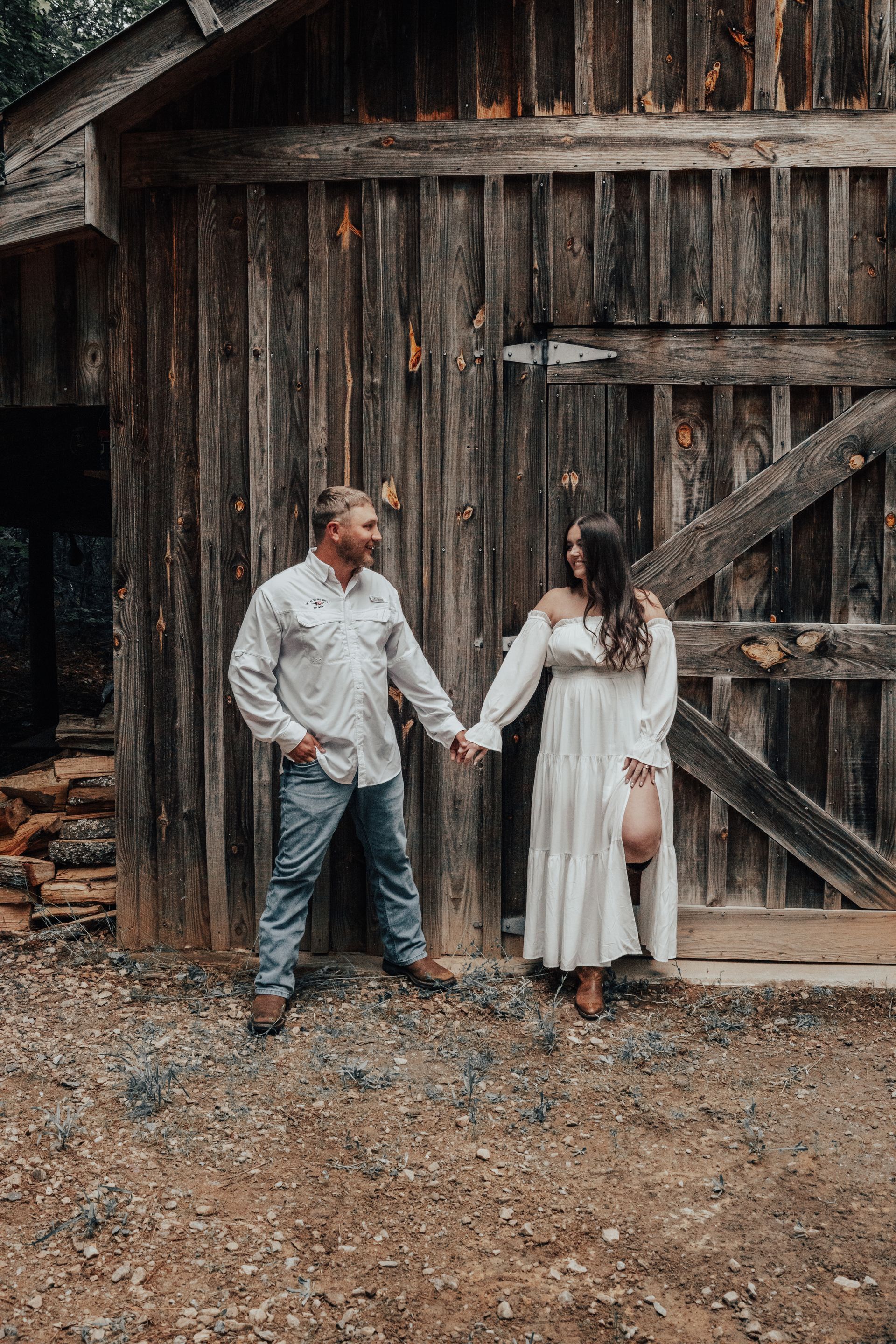 Couple holding hands in front of a rustic wooden barn. Man in jeans and white shirt, woman in white dress.