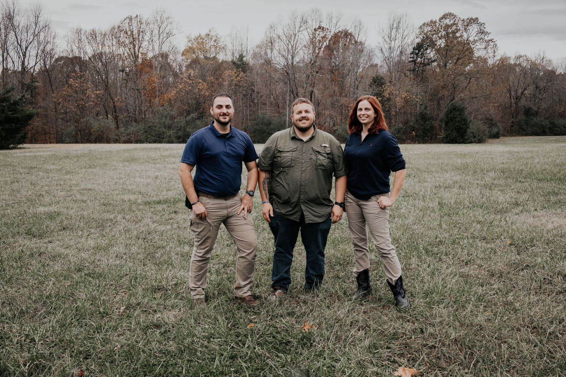 Three people standing in a grassy field with trees in the background. They are smiling, wearing casual clothing.