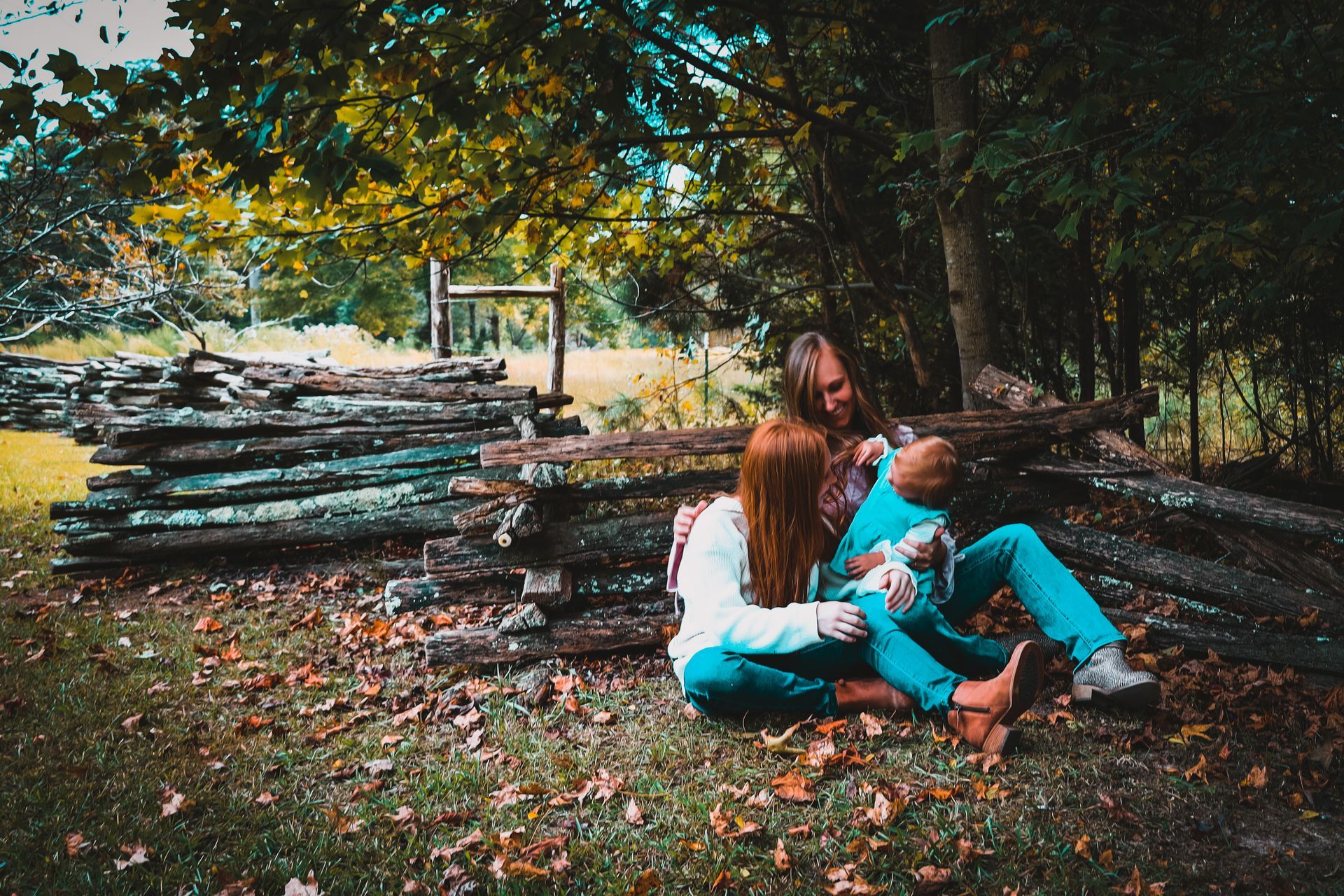Family sits together outside, holding a baby. They are in front of logs, under trees.