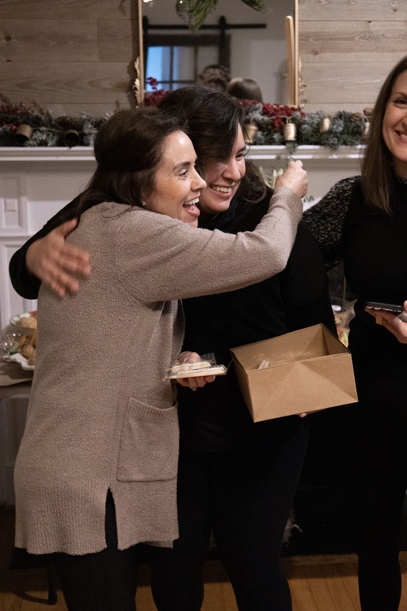 Woman in sweater hugs another, both smiling, holding box and plate with food. Indoors, festive decor.