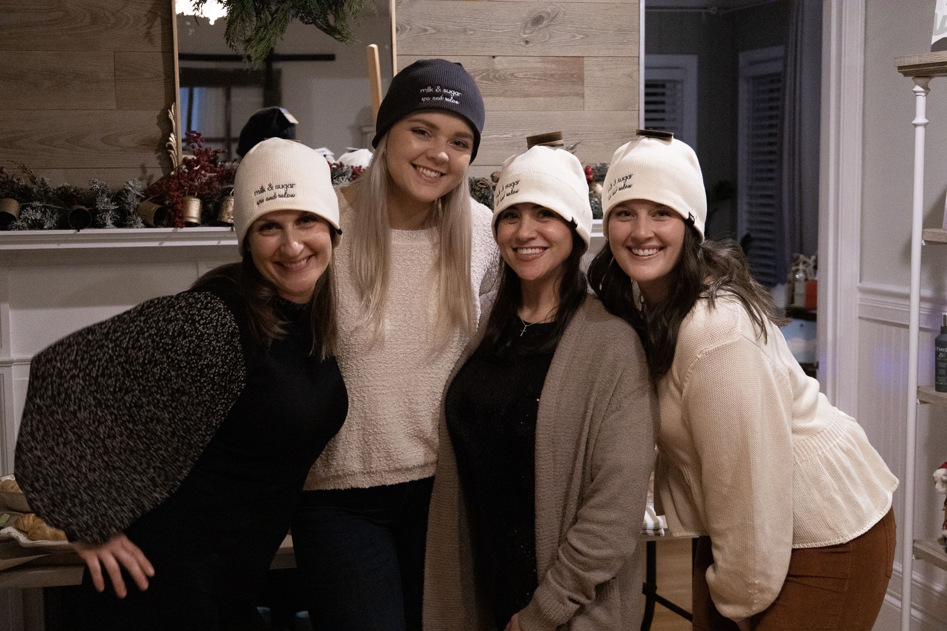 Four women smiling, wearing beanies and posing indoors.  One woman wears a dark hat; others, cream-colored.