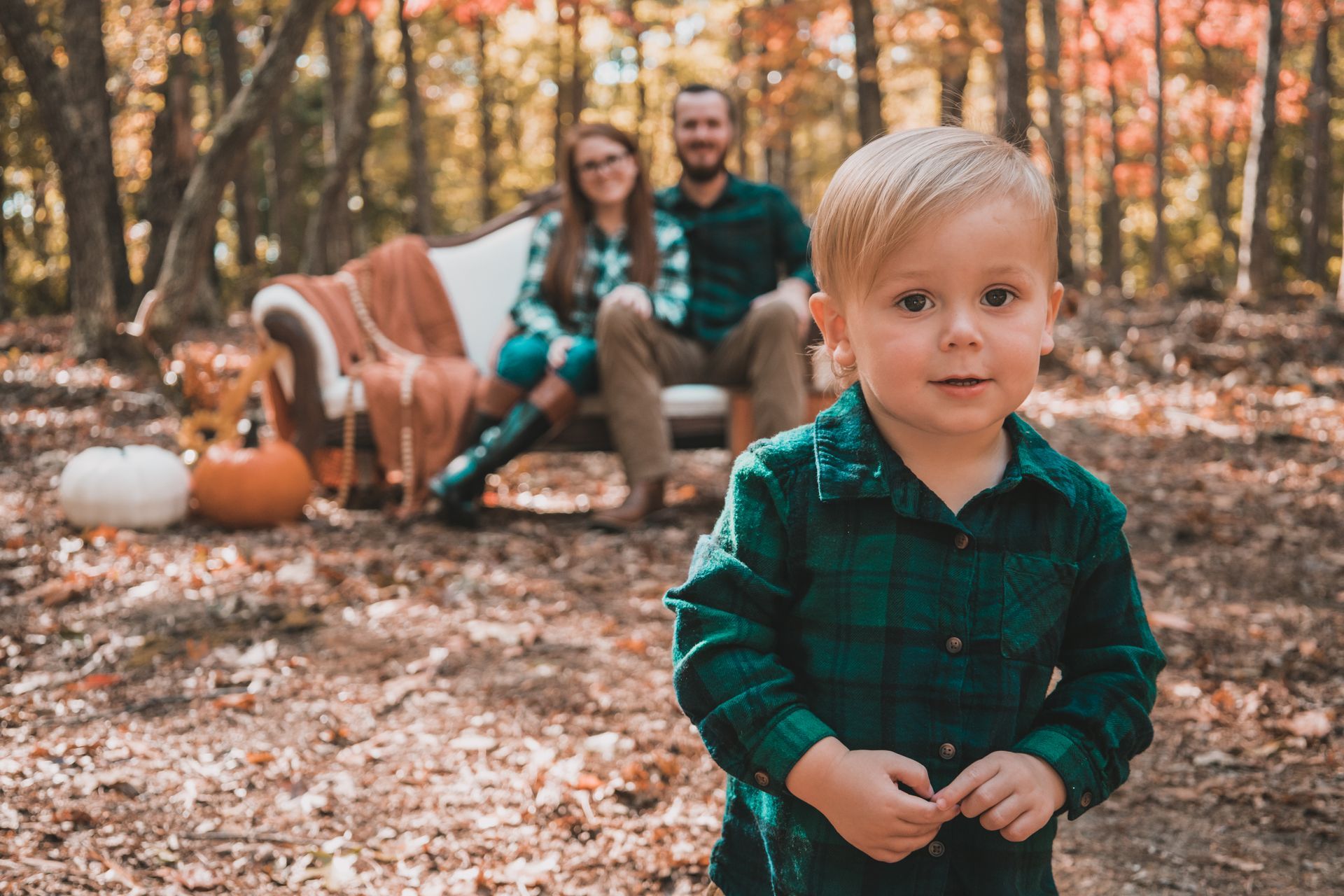Young child in green plaid shirt in focus; parents on a couch in the background in a fall forest setting.