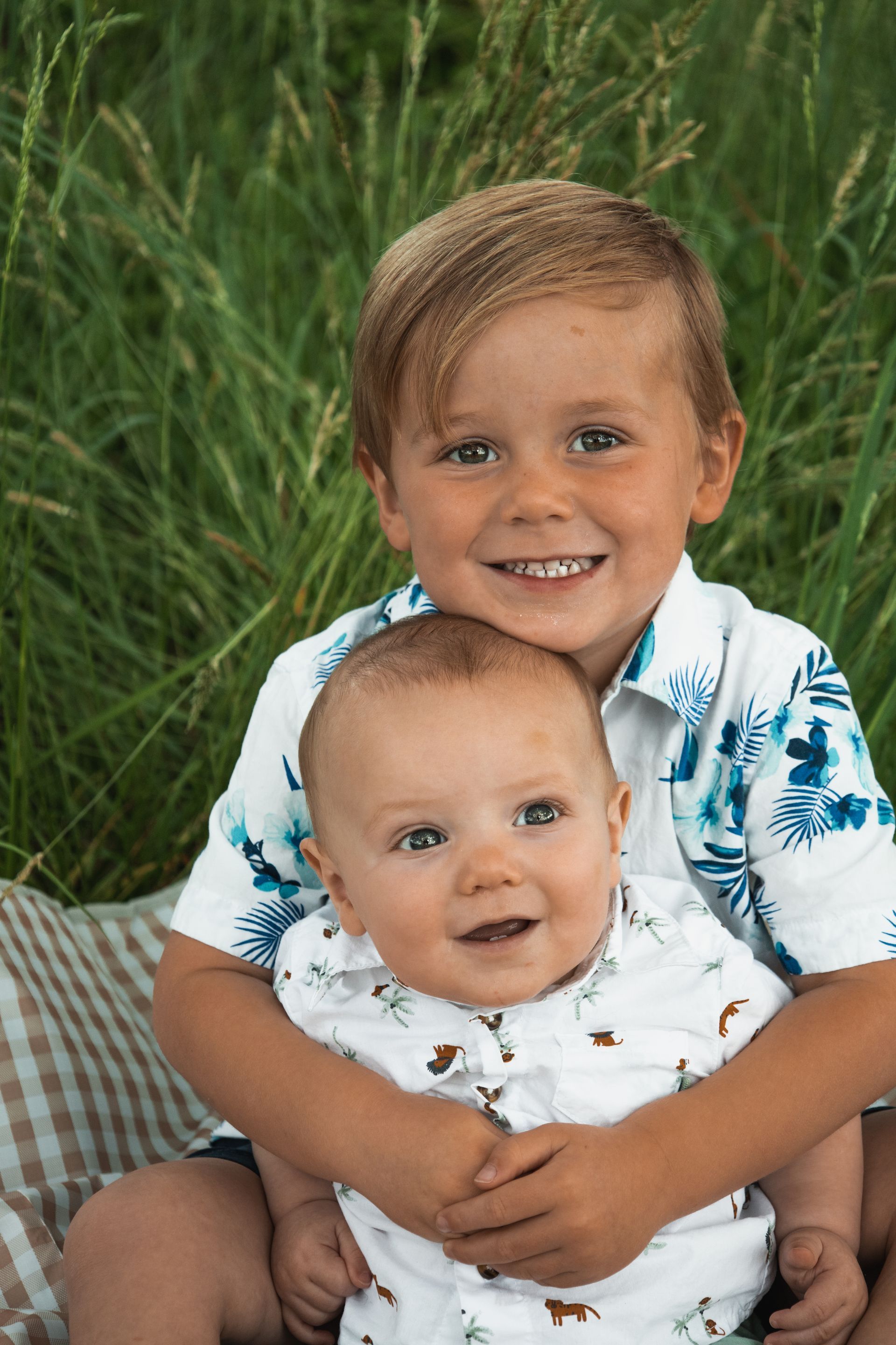Two young children smiling, one hugging the other in a grassy setting.
