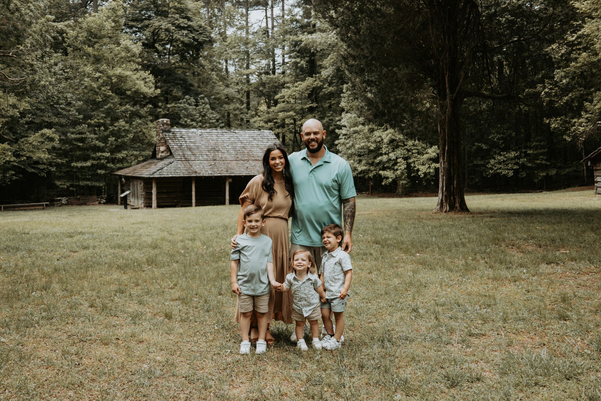 Family poses in front of a log cabin and trees: two parents, three children, wearing neutral tones.